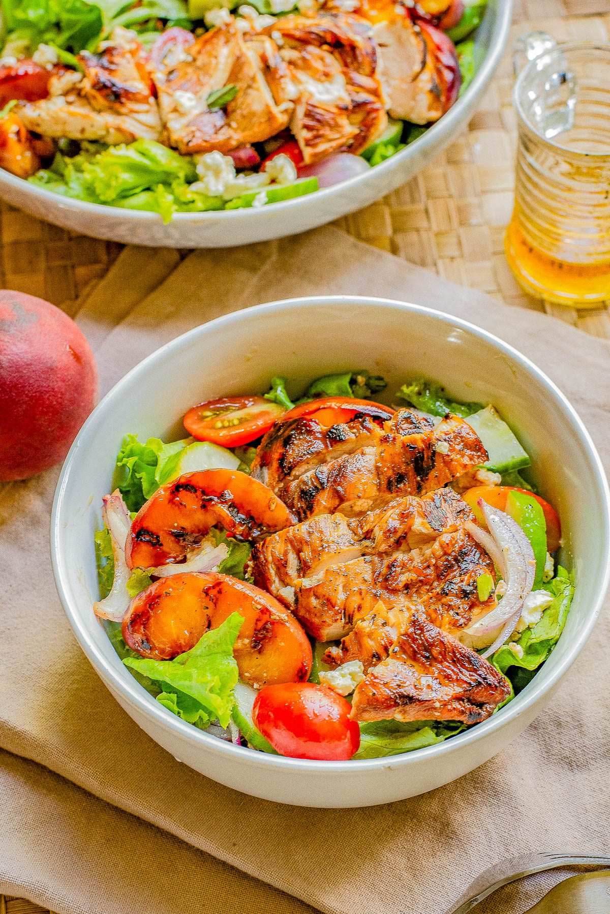 Two bowls of salad topped with grilled chicken, peaches, tomatoes, and leafy greens. One bowl is in the foreground, and a peach and a glass of iced tea can be seen on the table.