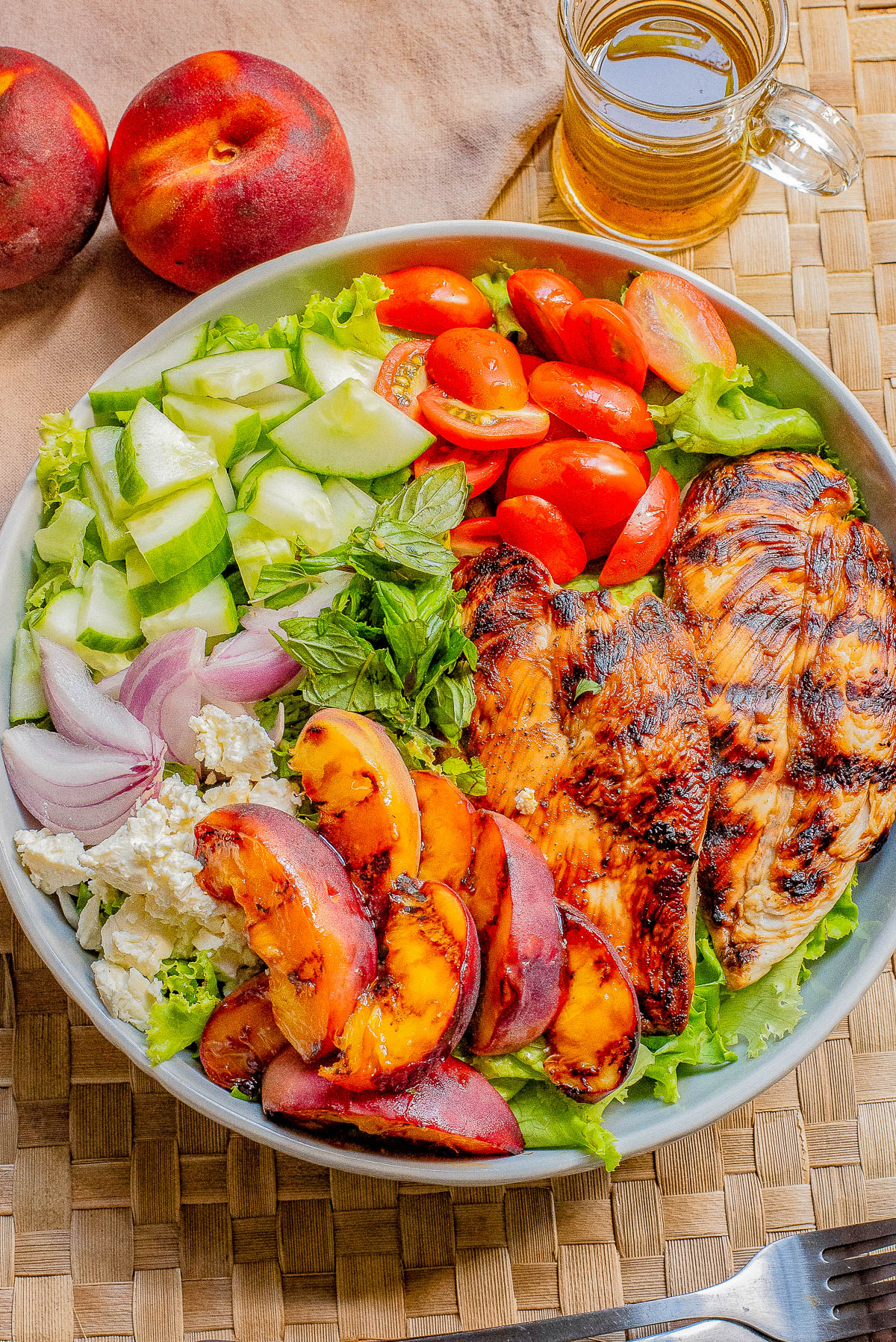 A bowl of salad with grilled chicken, grilled peaches, cherry tomatoes, cucumber, red onion, feta cheese, and herbs. Peaches and a mug of beverage are in the background.
