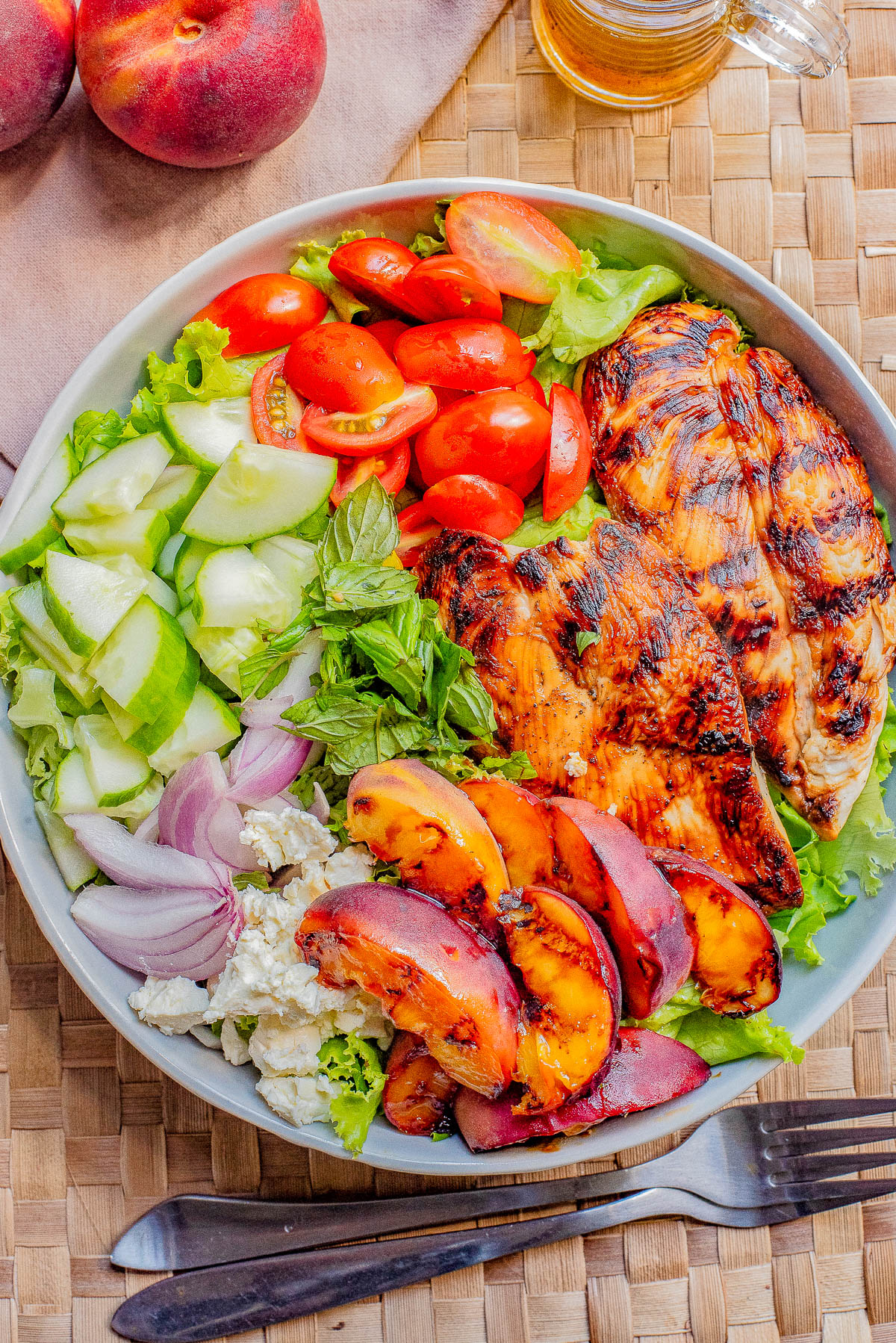 A salad bowl with grilled chicken, sliced peaches, cucumbers, cherry tomatoes, leafy greens, red onions, and feta cheese, placed on a woven mat with whole peaches and utensils beside it.