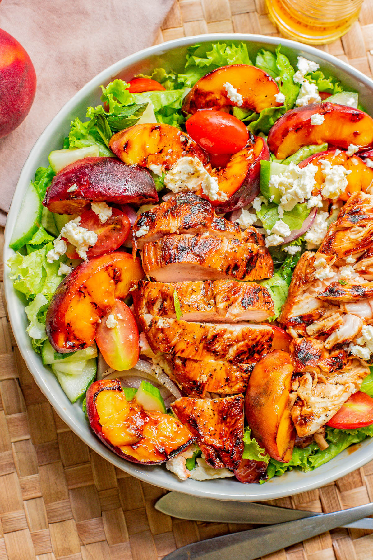 A bowl of salad featuring grilled chicken breast, peach slices, tomatoes, lettuce, and crumbled feta cheese, placed on a woven mat. A whole peach and a partial view of a glass bottle are in the background.