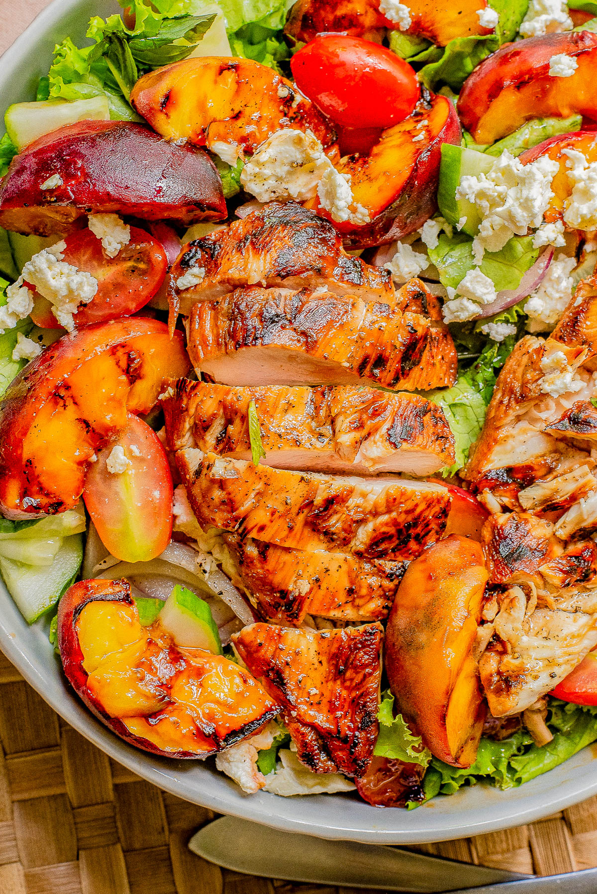 A healthy salad featuring grilled chicken, assorted greens, tomatoes, peaches, and crumbled cheese, served in a bowl on a woven mat.