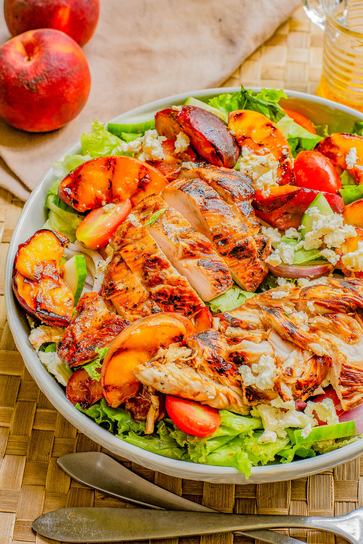 A bowl of grilled chicken salad with peaches, tomatoes, lettuce, and crumbled cheese, placed on a woven placemat with peaches and a drink in the background.