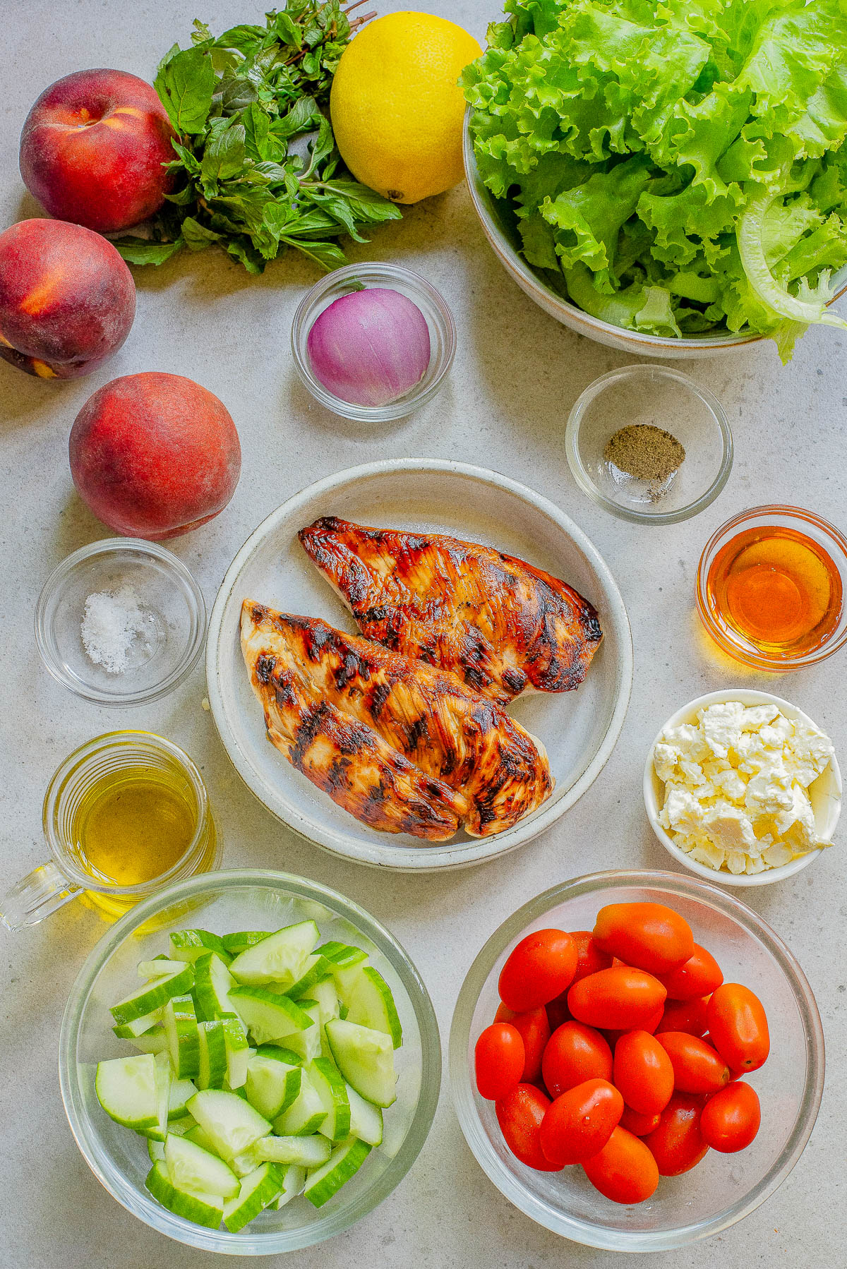 A variety of fresh ingredients on a table, including lettuce, peaches, herbs, lemon, red onion, grilled chicken, cucumber slices, cherry tomatoes, feta cheese, olive oil, pepper, salt, and vinegar.