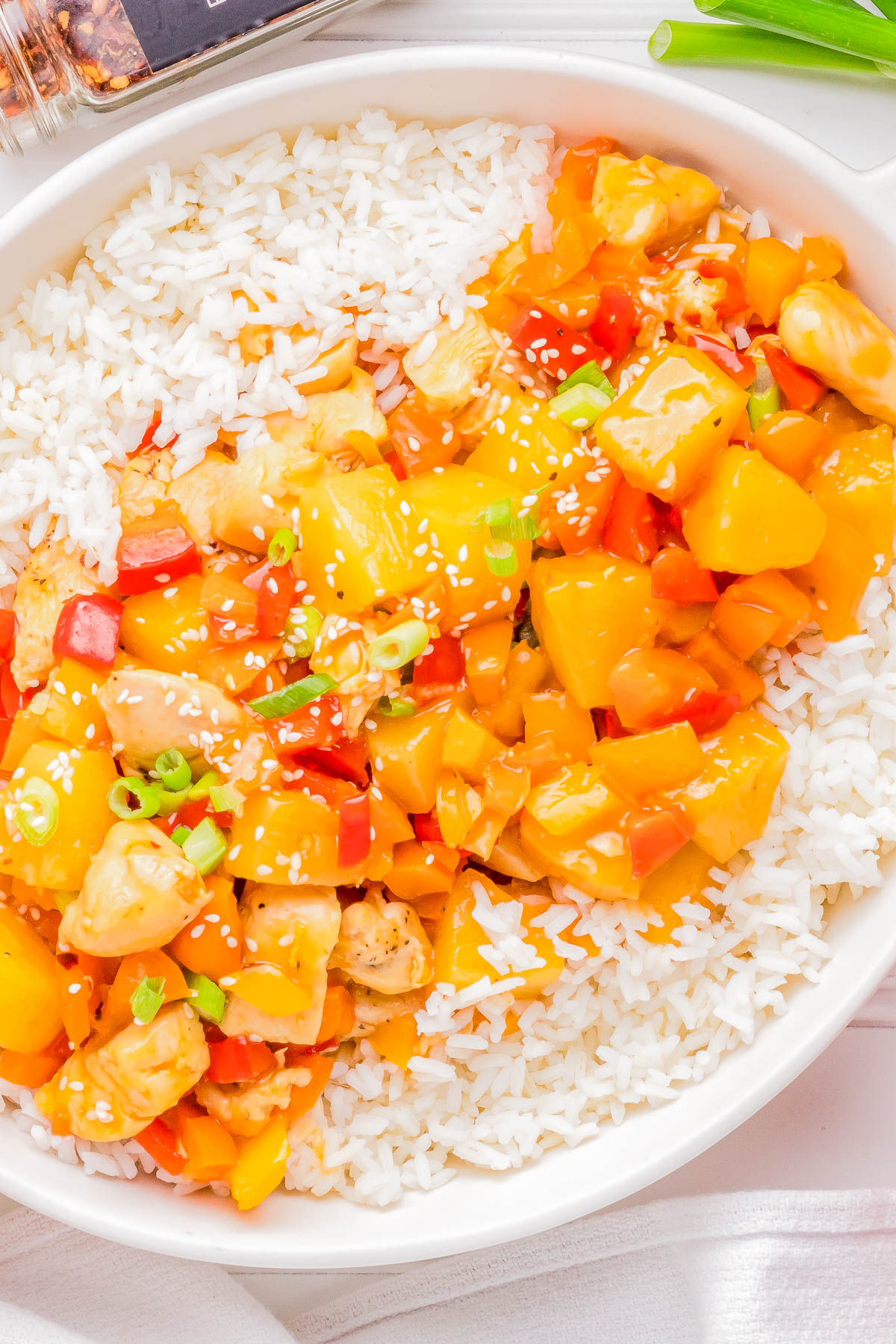 A bowl of white rice topped with diced chicken, red bell peppers, pineapples, and sesame seeds, served with orange-colored sauce.