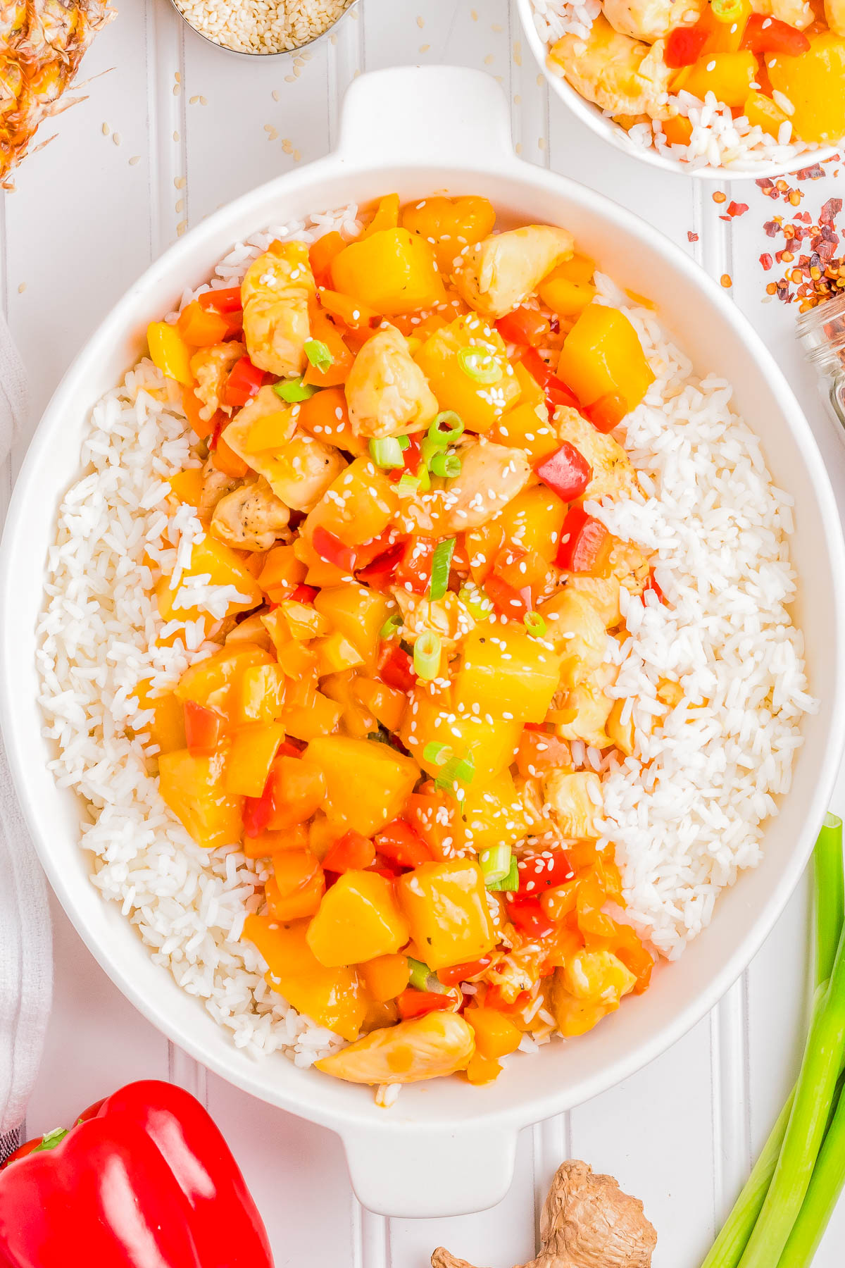 A white bowl filled with rice topped with a colorful mix of diced chicken, pineapple, bell peppers, and sesame seeds. Various ingredients and seasonings are visible in the background.