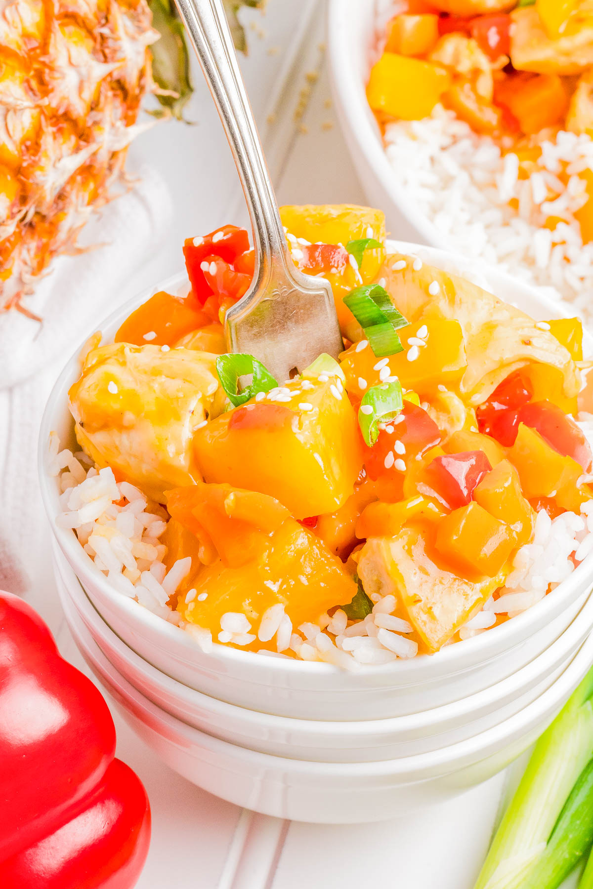 A bowl of rice topped with colorful sweet and sour chicken garnished with green onions and sesame seeds. A fork is placed in the bowl, and a red bell pepper and pineapple are partially visible.