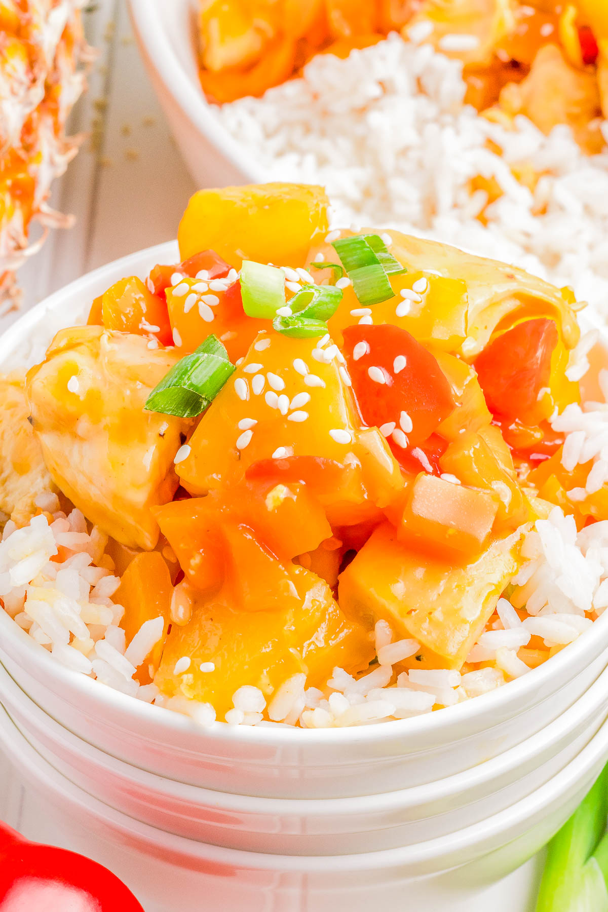 A close-up of a white bowl filled with rice, topped with chunks of pineapple, bell peppers, and tofu in a bright sauce, garnished with sesame seeds and chopped green onions.