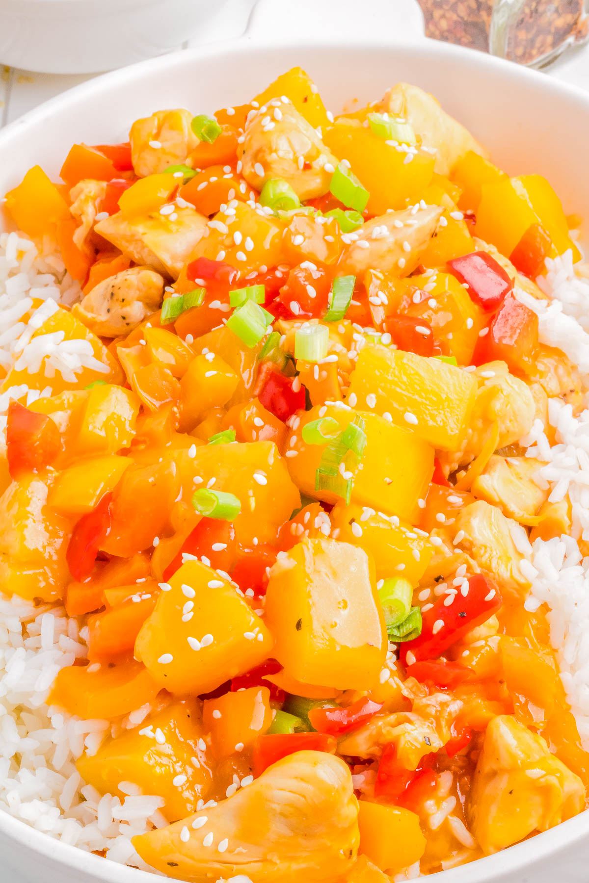 A bowl of white rice topped with pieces of chicken, bell peppers, pineapple chunks, and a glossy orange sauce, garnished with sesame seeds and chopped green onions.