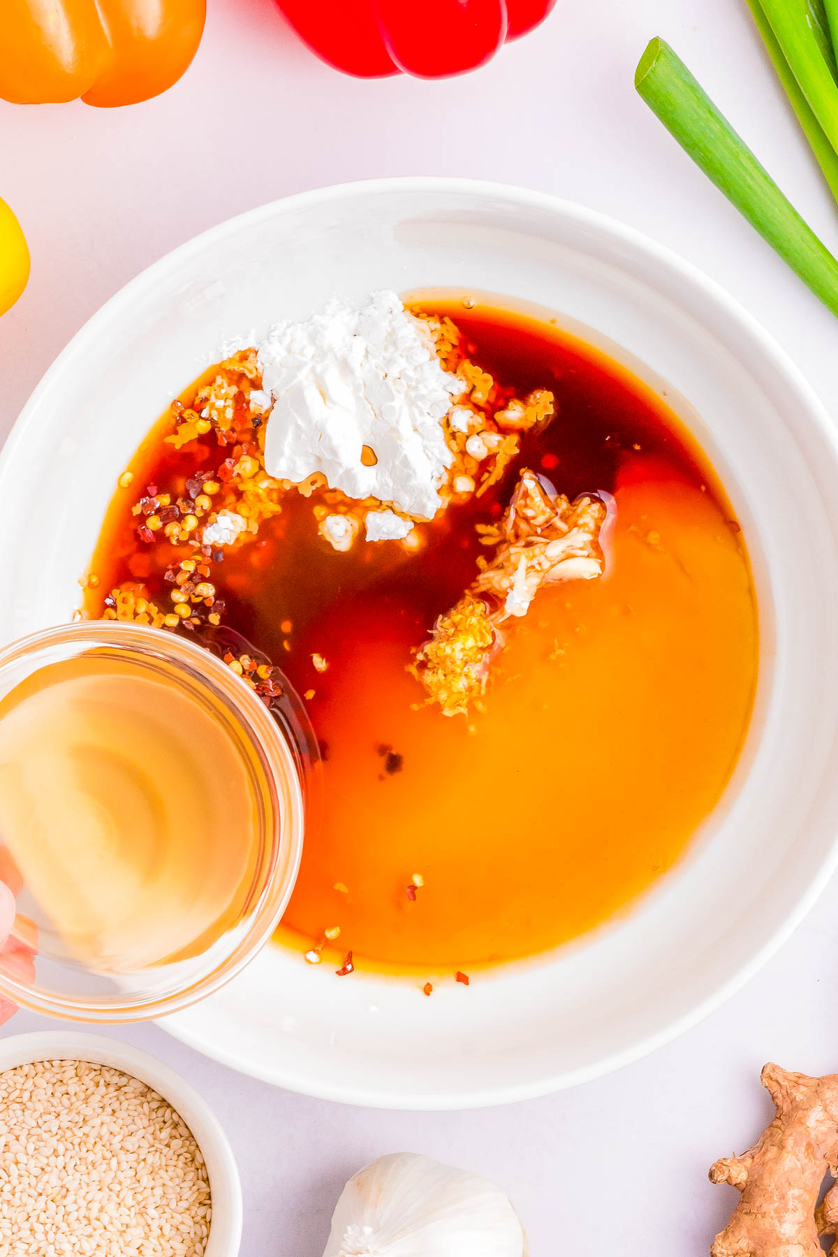 A shallow white bowl contains a mix of dark and light orange sauces with white powder and a sprinkle of seeds. A hand is pouring a light yellow liquid into the bowl. Nearby are garlic, sesame, and green onions.