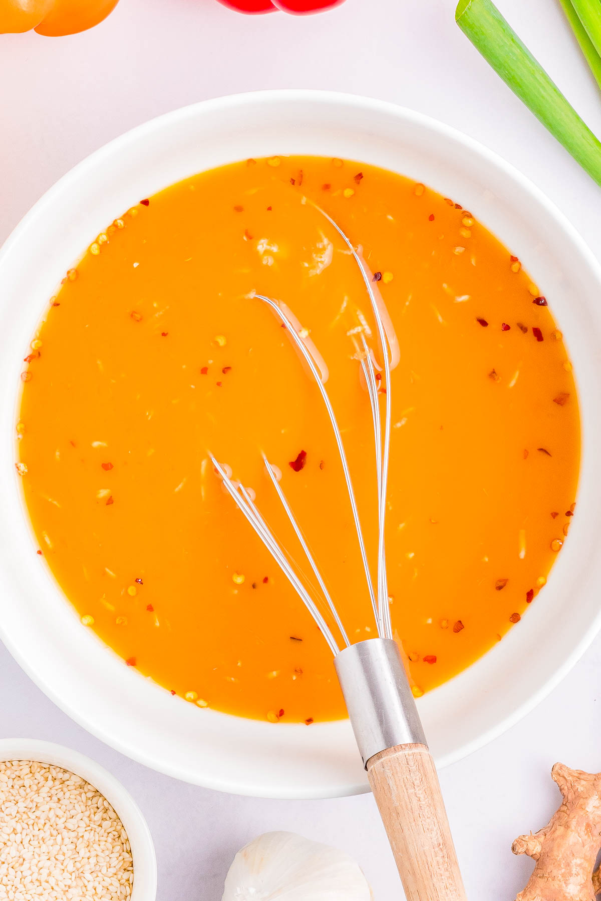 A bowl filled with orange-colored sauce garnished with red pepper flakes and seasonings, with a whisk resting inside. Scallions, garlic, sesame seeds, and ginger are visible around the bowl.