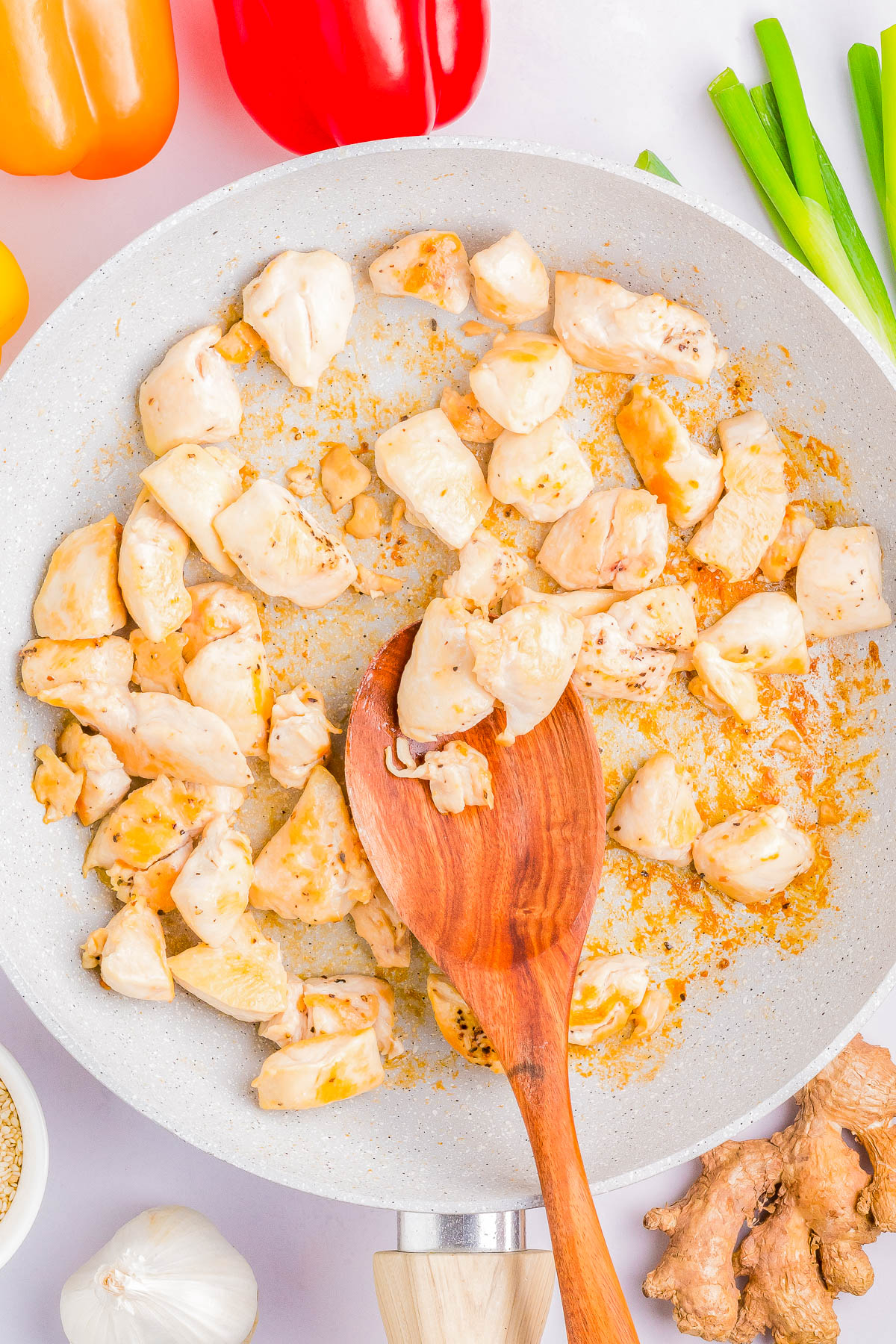 Diced chicken pieces being cooked in a frying pan with a wooden spoon. Various ingredients, including bell peppers, green onions, garlic, ginger, and sesame seeds, surround the pan.