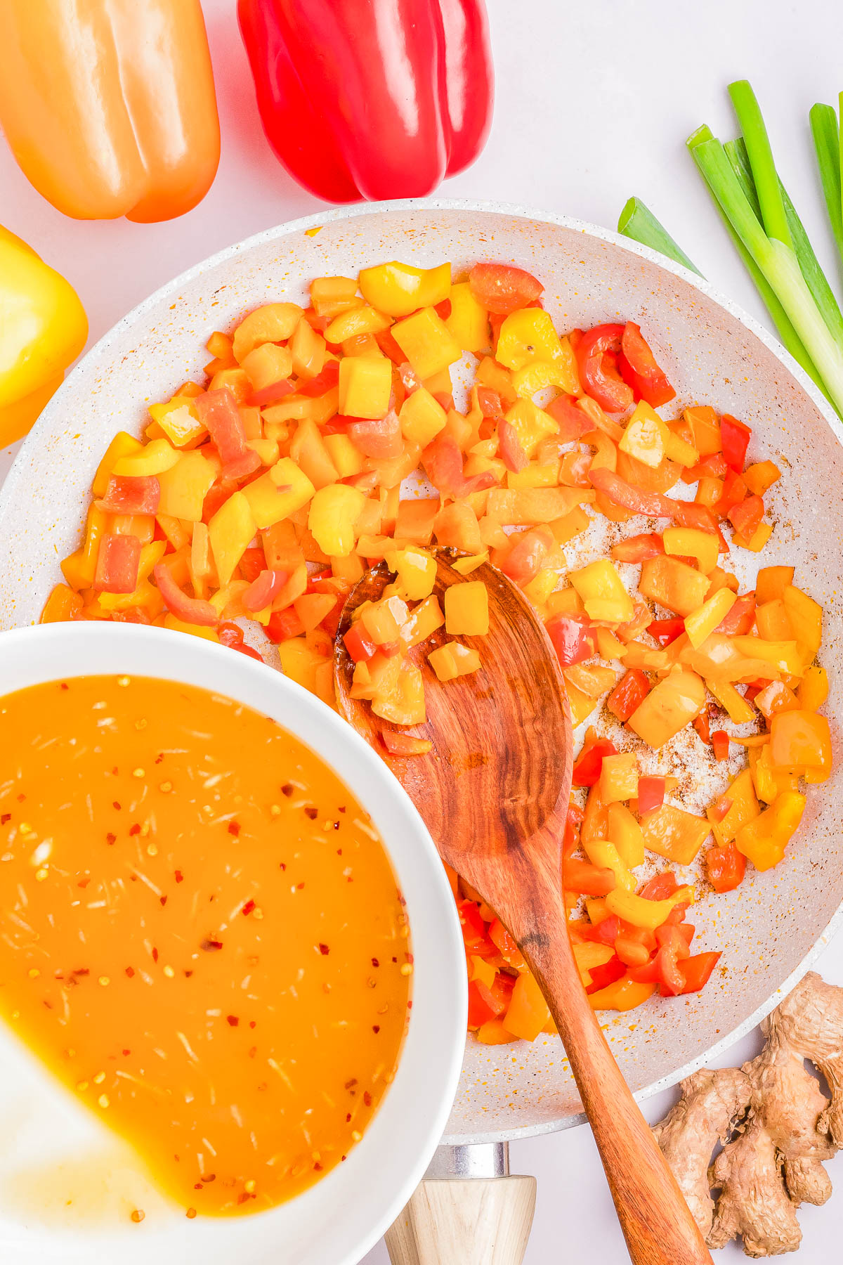 Chopped red and yellow bell peppers being sautéed in a pan with a wooden spoon. A bowl of chili sauce, sliced green onions, and fresh ginger are also visible nearby.