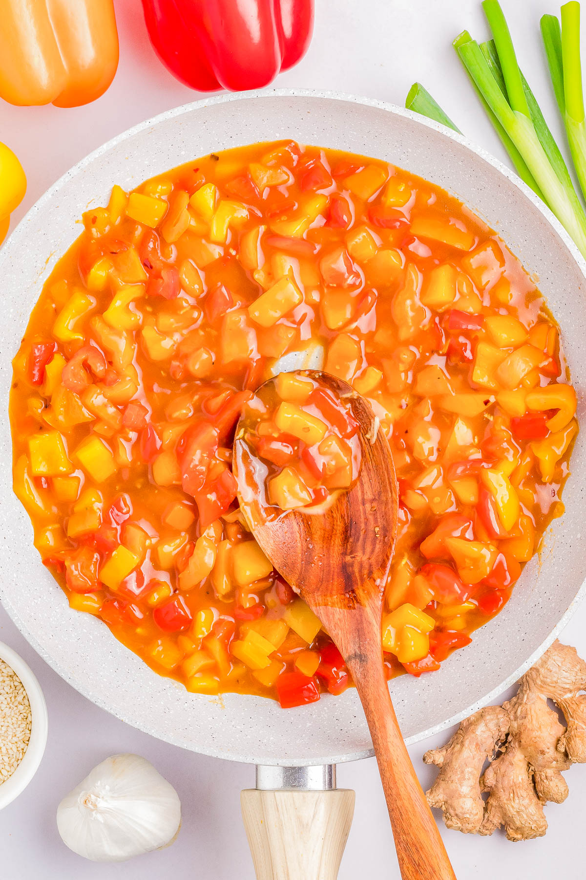 A pan filled with a vibrant, saucy mixture of chopped yellow and red bell peppers, with a wooden spoon stirring it. Fresh vegetables like green onions, ginger, and garlic surround the pan.