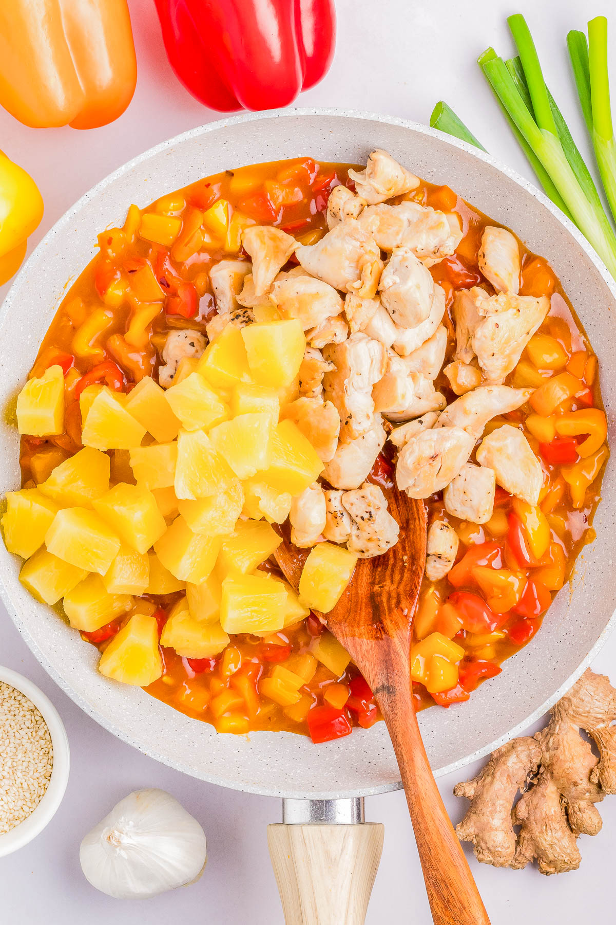 A pan with diced chicken, pineapple chunks, and a colorful mixture of red, yellow, and orange bell peppers. Green onions, garlic, ginger, sesame seeds, and bell peppers are arranged around the pan.