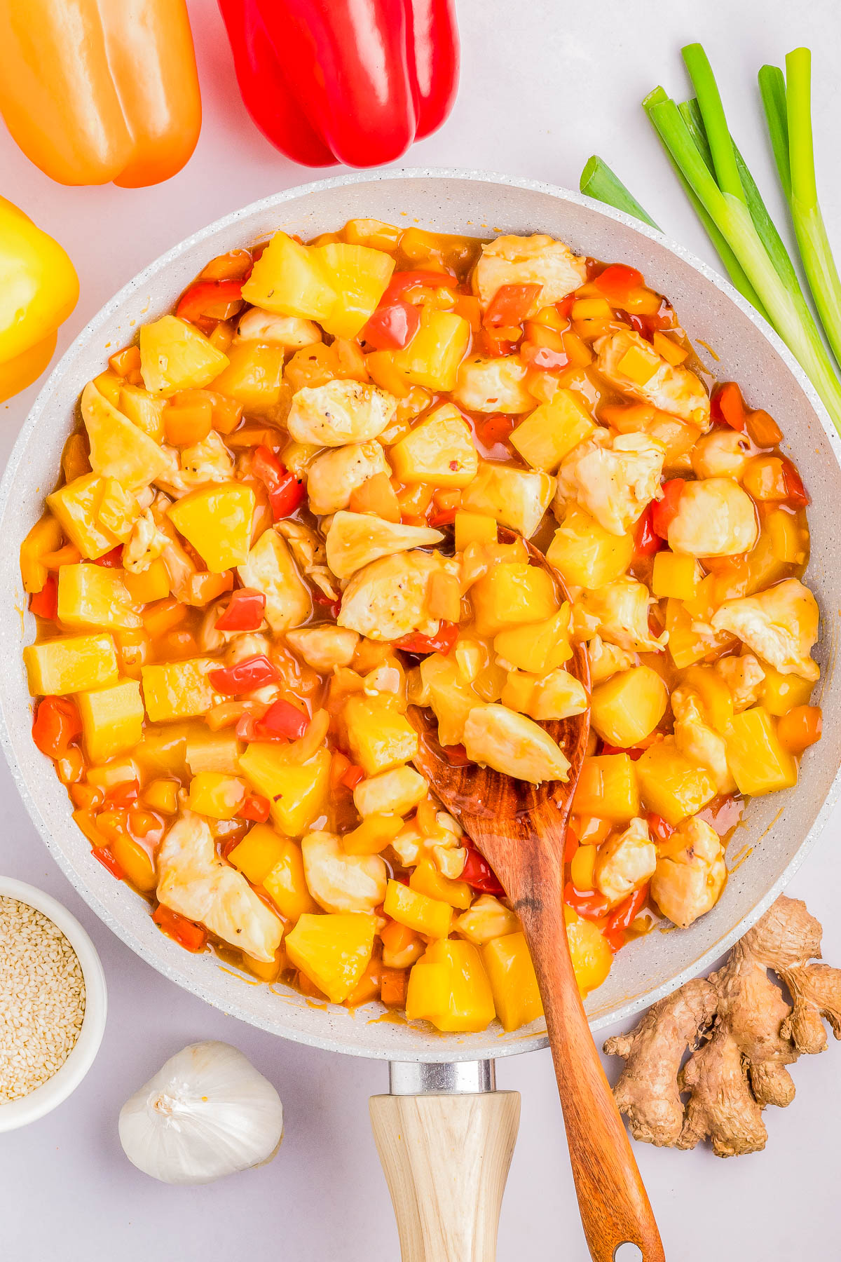 A skillet filled with sweet and sour chicken with pineapple chunks and bell peppers, surrounded by ingredients including ginger, garlic, green onions, and sesame seeds.