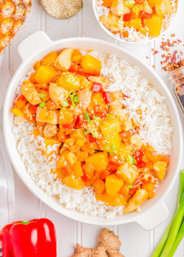 A dish of sweet and sour chicken with pineapple and bell peppers served over white rice in a white bowl. Nearby, a smaller bowl with the same dish, a red bell pepper, fresh ginger, and sesame seeds.