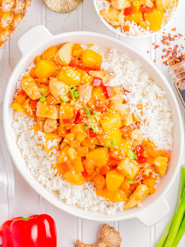 A dish of sweet and sour chicken with pineapple and bell peppers served over white rice in a white bowl. Nearby, a smaller bowl with the same dish, a red bell pepper, fresh ginger, and sesame seeds.