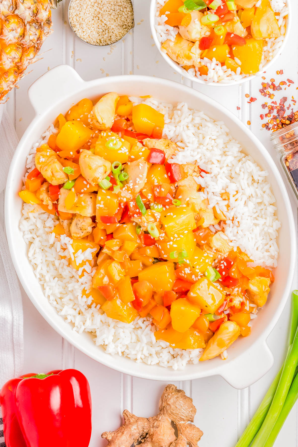 A dish of sweet and sour chicken with pineapple and bell peppers served over white rice in a white bowl. Nearby, a smaller bowl with the same dish, a red bell pepper, fresh ginger, and sesame seeds.