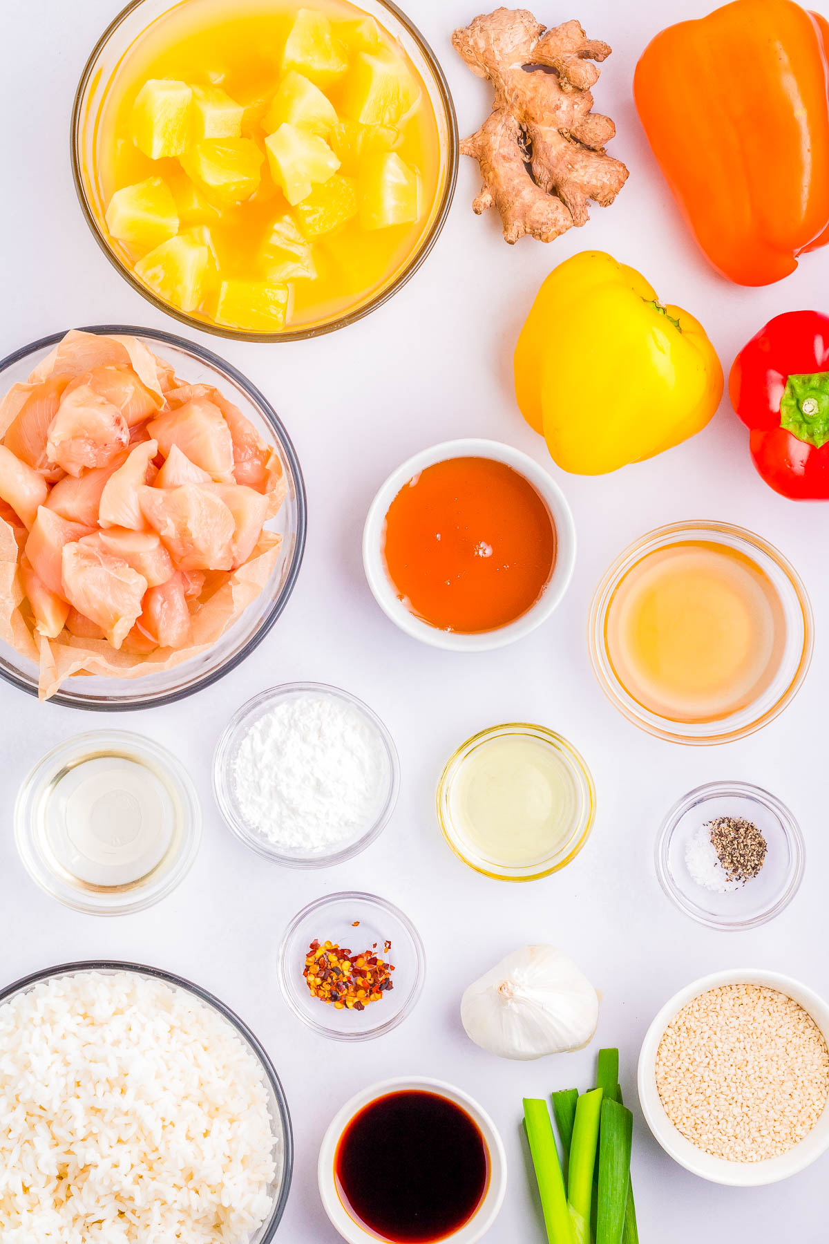 Top view of various ingredients laid out for a sweet and sour chicken recipe, including diced chicken, pineapples, ginger, bell peppers, rice, seasonings, sauces, and spices on a white surface.