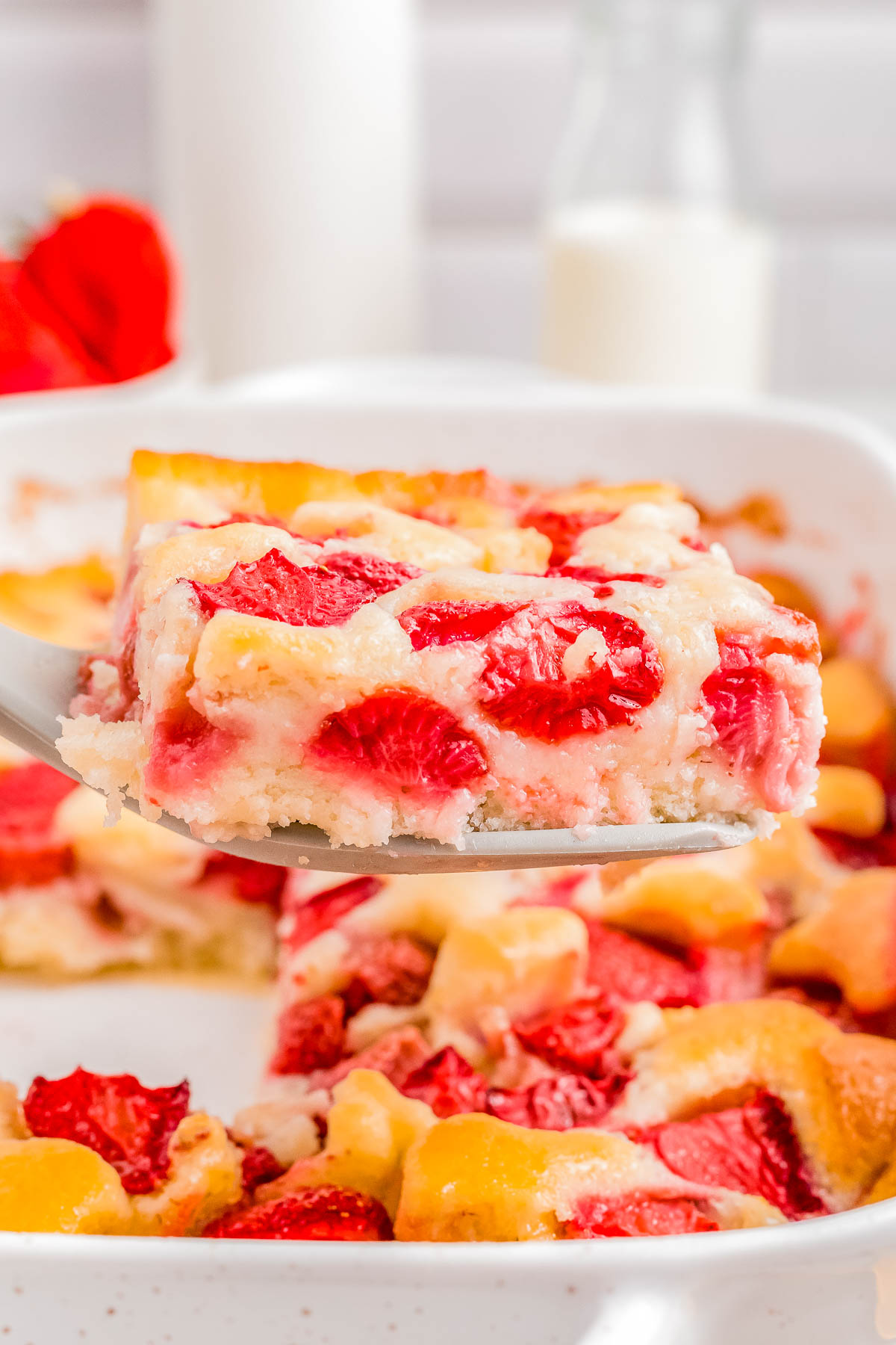 A slice of strawberry cobbler being lifted from a dish, showcasing ripe strawberries and a fluffy texture, with a glass of milk in the background.