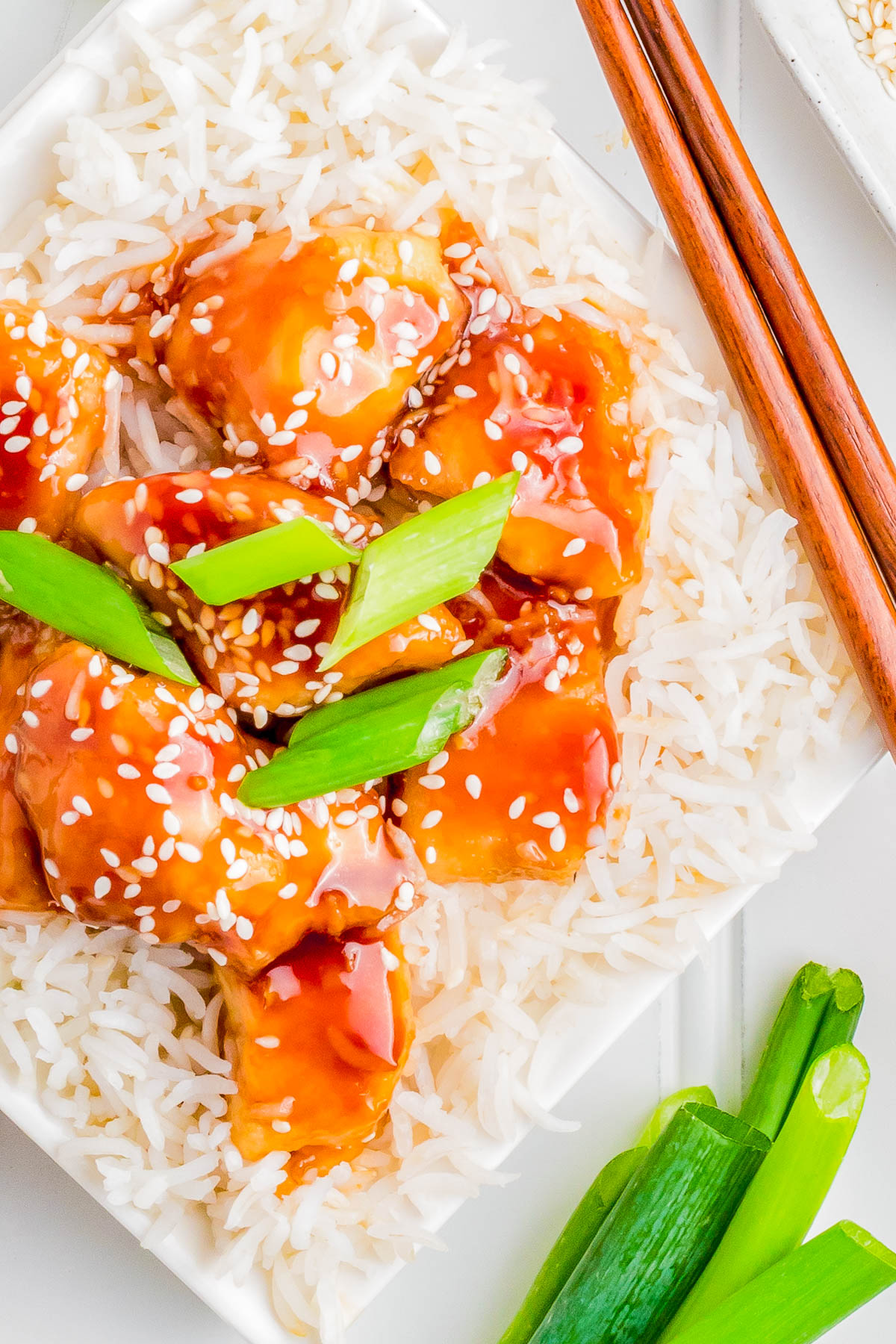 A plate of white rice topped with pieces of chicken glazed in an sauce garnished with white sesame seeds and sliced green onions. Chopsticks rest on the side.