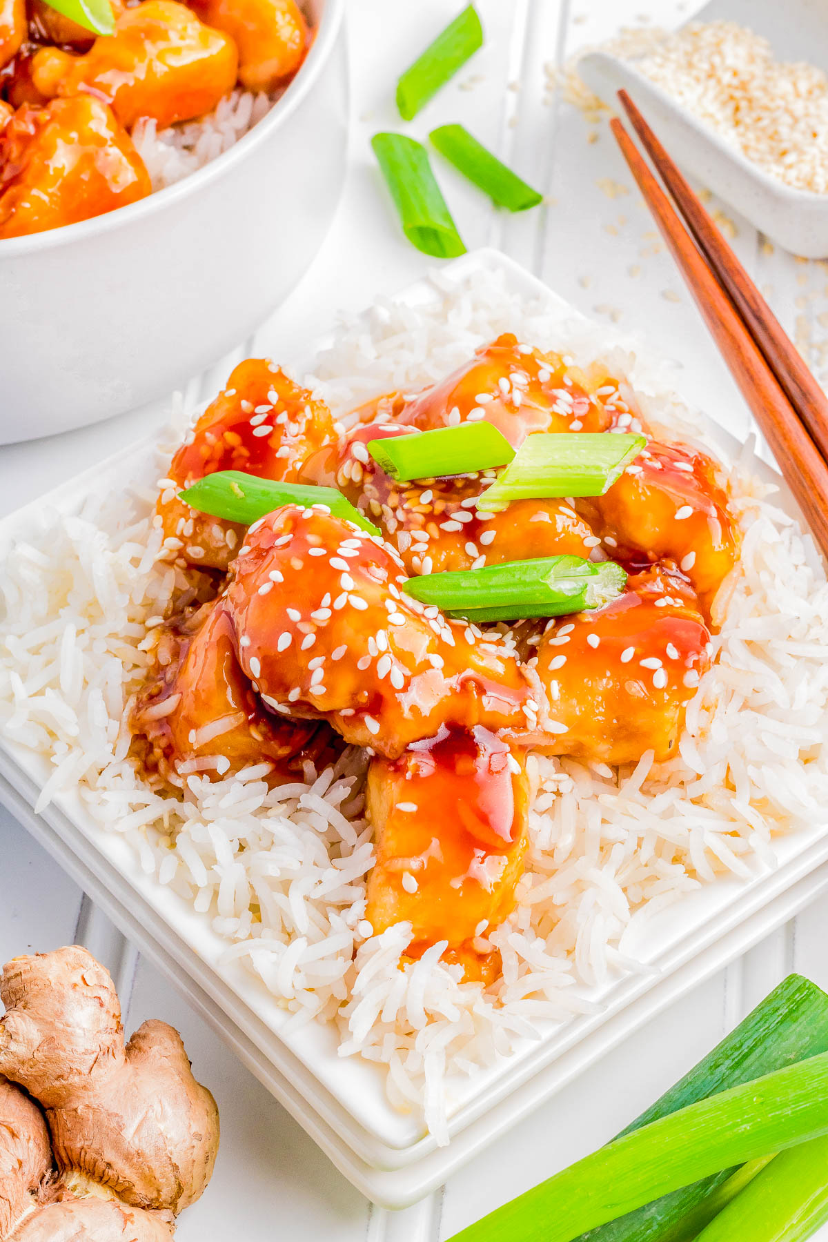 A square white plate of rice topped with sauced chicken chunks, garnished with sesame seeds and green onions, with a pair of wooden chopsticks lying beside the plate.