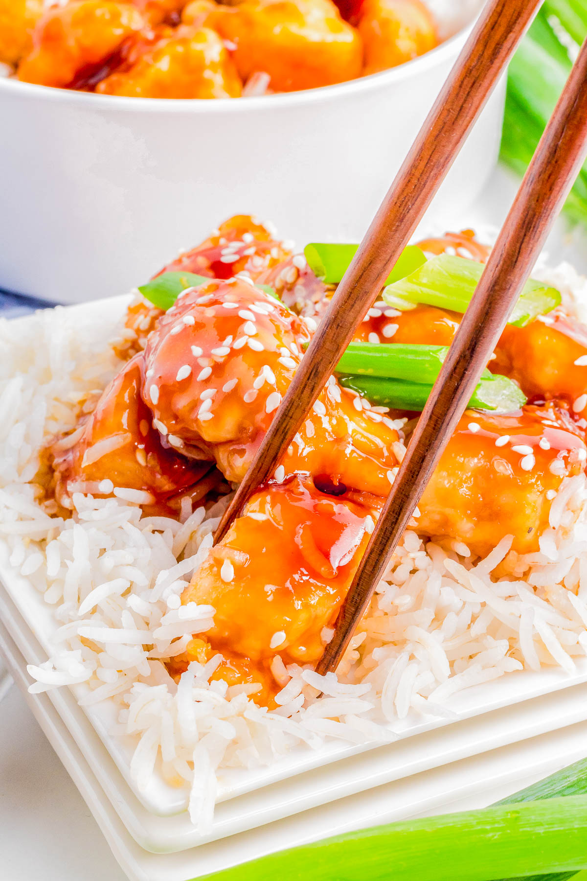 A close-up of chopsticks holding a piece of Mongolian chicken over a bed of white rice, garnished with sesame seeds and green onions.