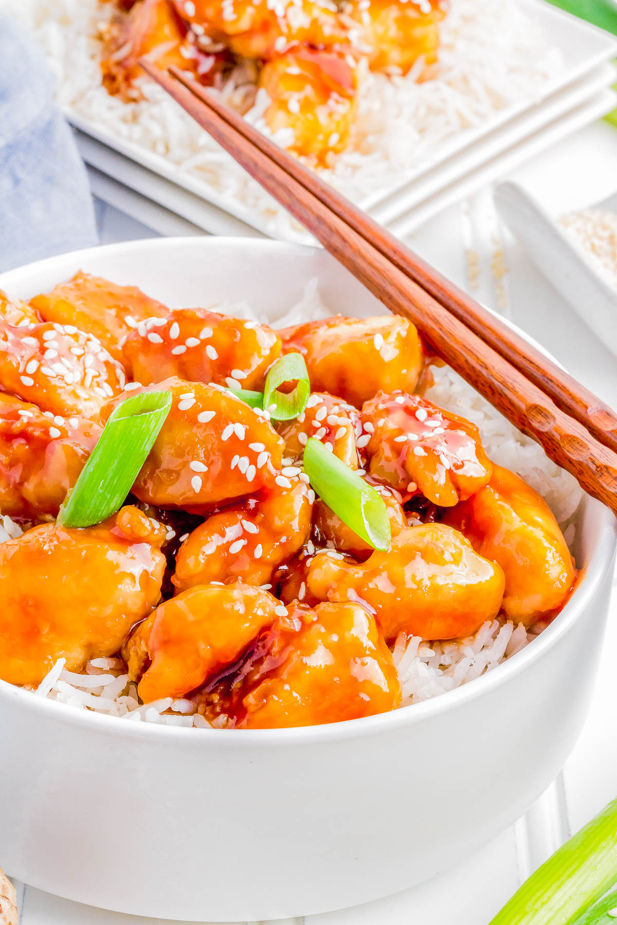 A bowl of sesame chicken garnished with green onions and sesame seeds, served over white rice with chopsticks resting on the edge.