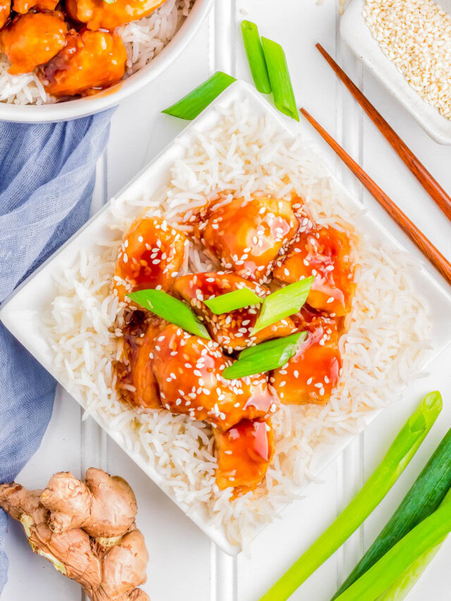 A square white plate with rice topped with glazed chicken chunks, garnished with sesame seeds and green onions. Chopsticks, fresh ginger, green onions, and a napkin are beside the plate.