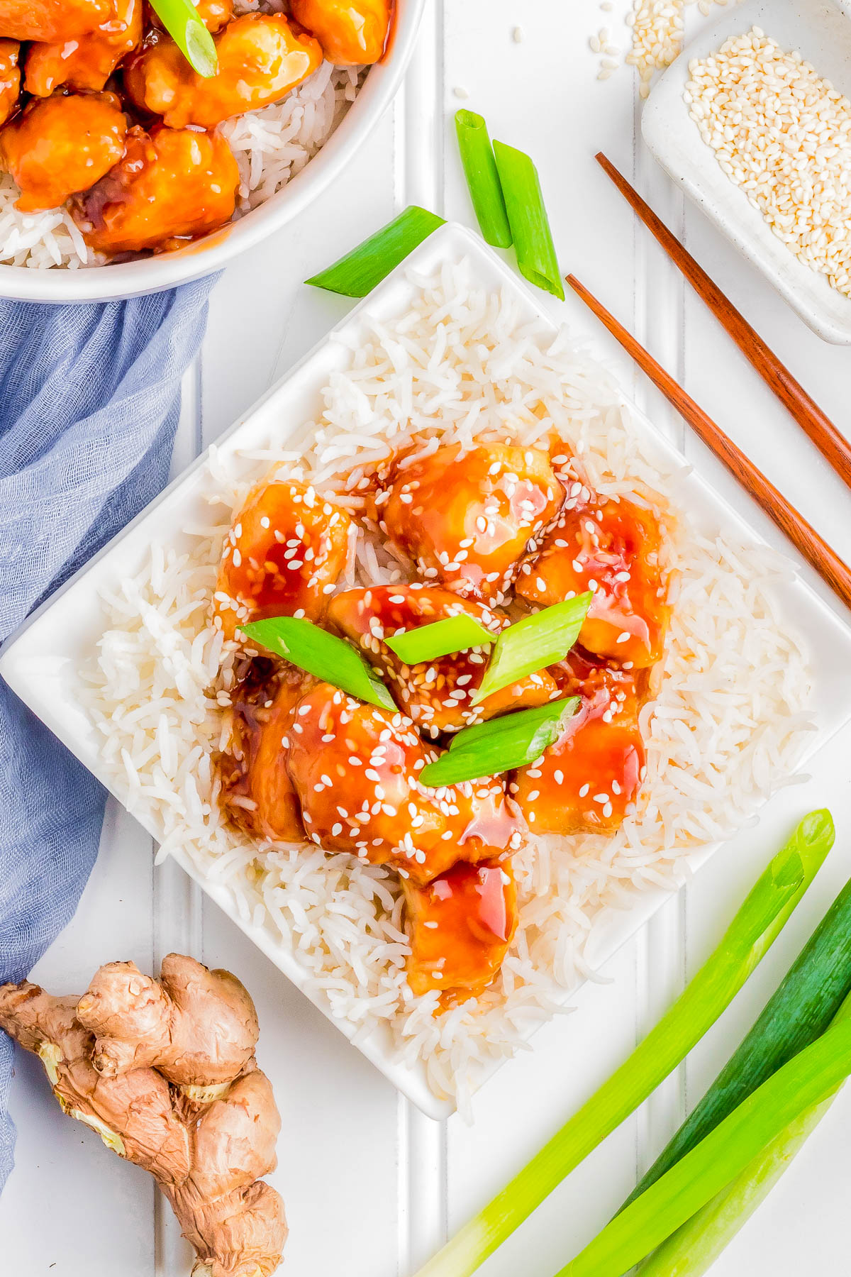 A square white plate with rice topped with glazed chicken chunks, garnished with sesame seeds and green onions. Chopsticks, fresh ginger, green onions, and a napkin are beside the plate.