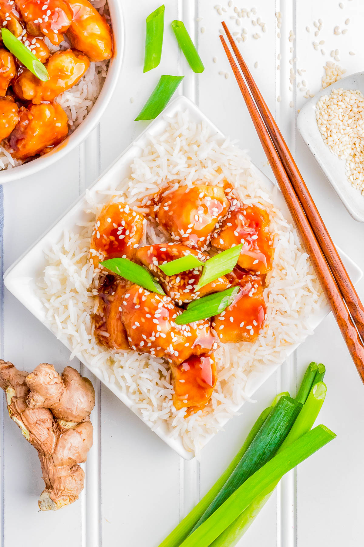 A plate of white rice topped with glazed chicken pieces garnished with sesame seeds and green onions. Fresh ginger, green onions, sesame seeds, and a bowl of chicken are visible nearby.