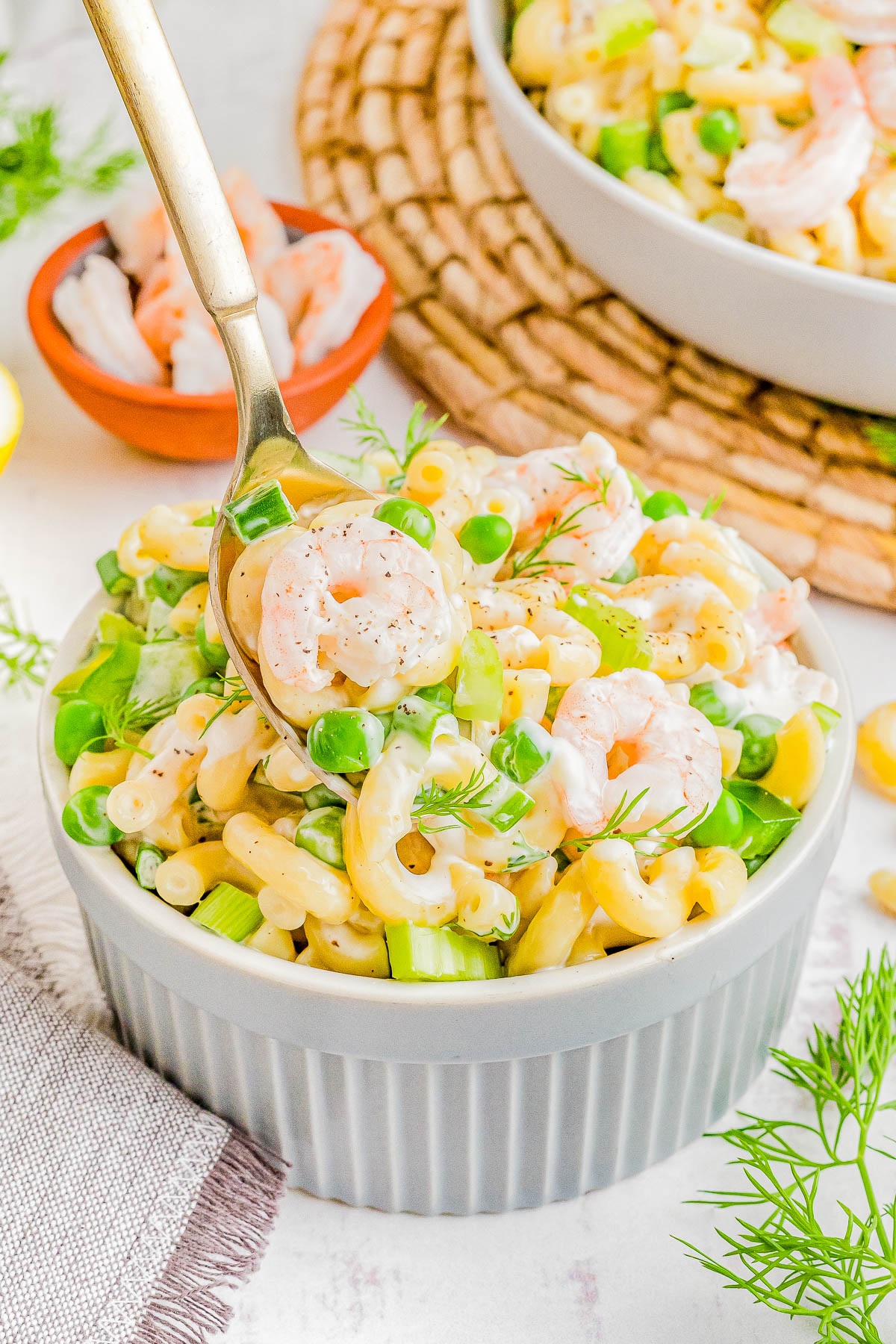 A bowl of creamy shrimp pasta salad garnished with fresh dill and peas, served on a light table with crackers in the background.