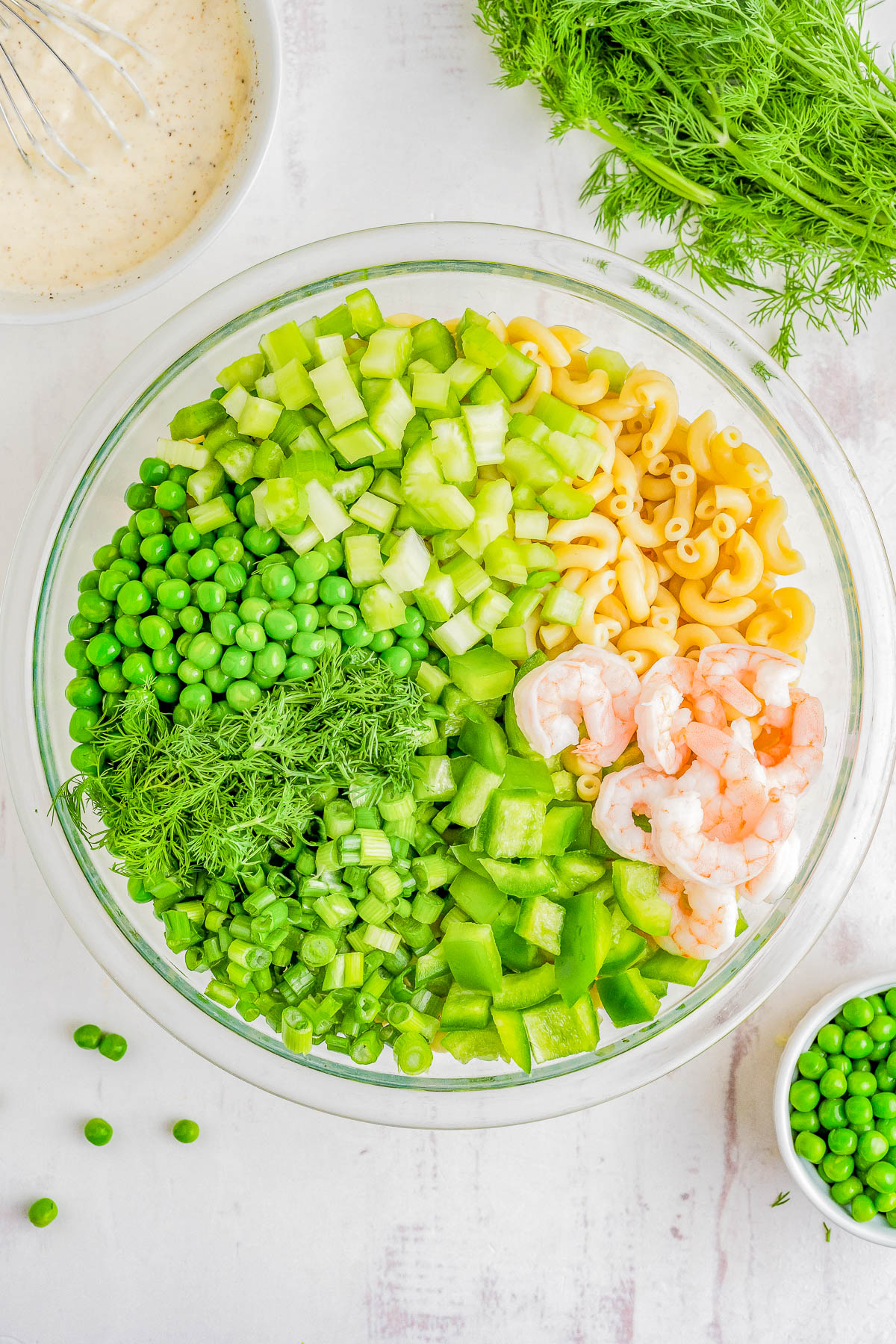 A glass bowl containing a salad with pasta, shrimp, green peas, chopped cucumber, and fresh dill, with a dressing on the side.