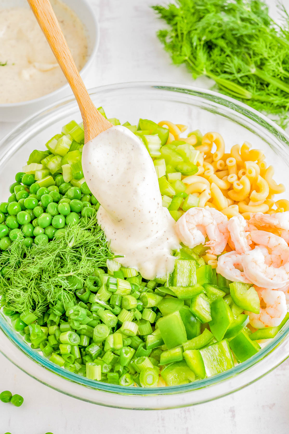 A glass bowl contains peas, green beans, dill, elbow pasta, and shrimp, with dressing being poured over it.