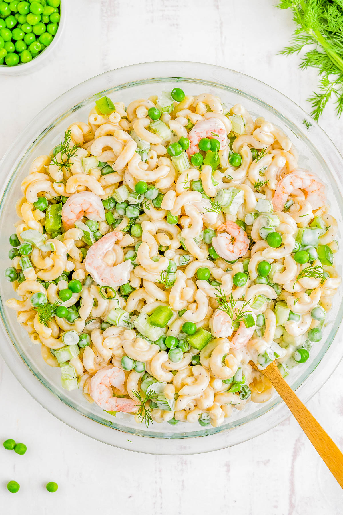 A bowl of shrimp pasta salad with peas, celery, and fresh dill, served on a white background with a wooden spoon.