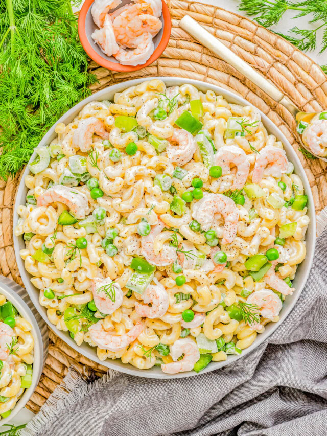 A vibrant shrimp pasta salad with green peas, dill, and cashews, served on a white platter over a rustic wicker mat and grey linen.