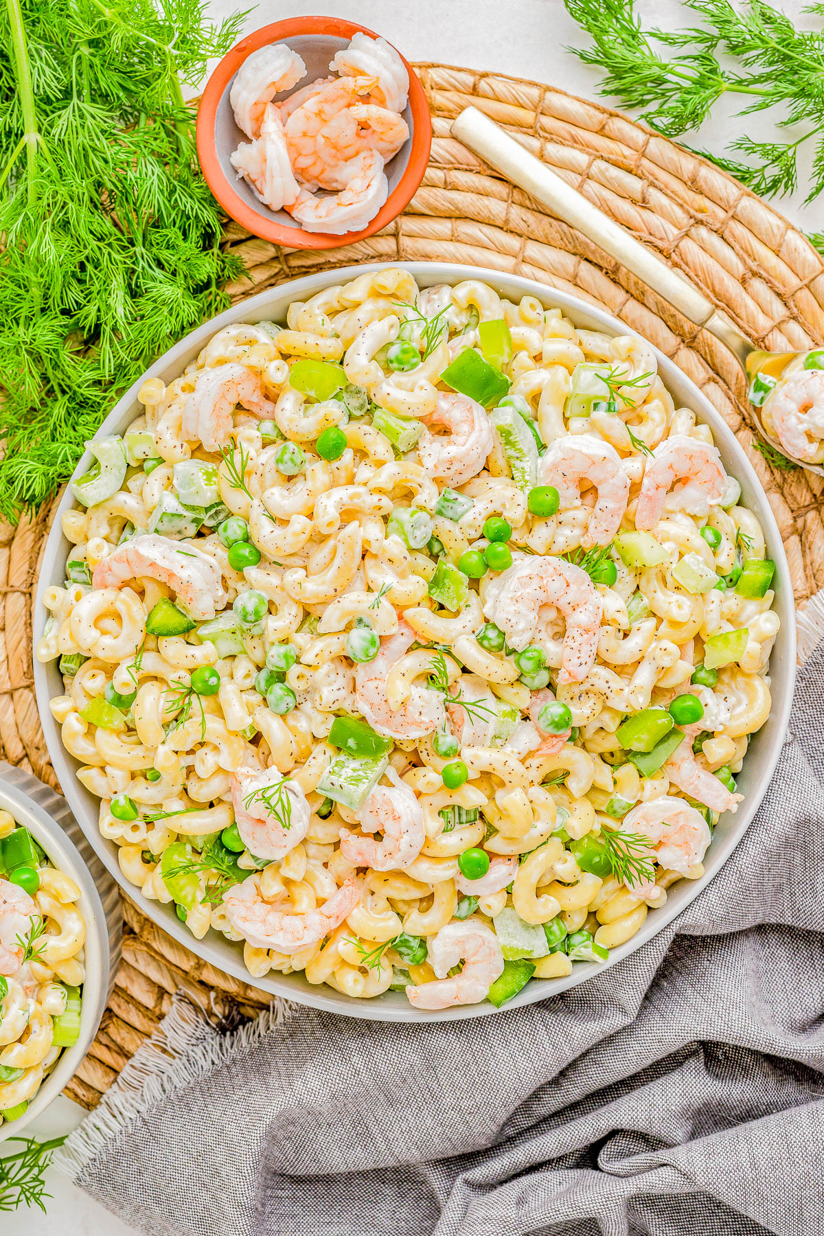 A vibrant shrimp pasta salad with green peas, dill, and cashews, served on a white platter over a rustic wicker mat and grey linen.