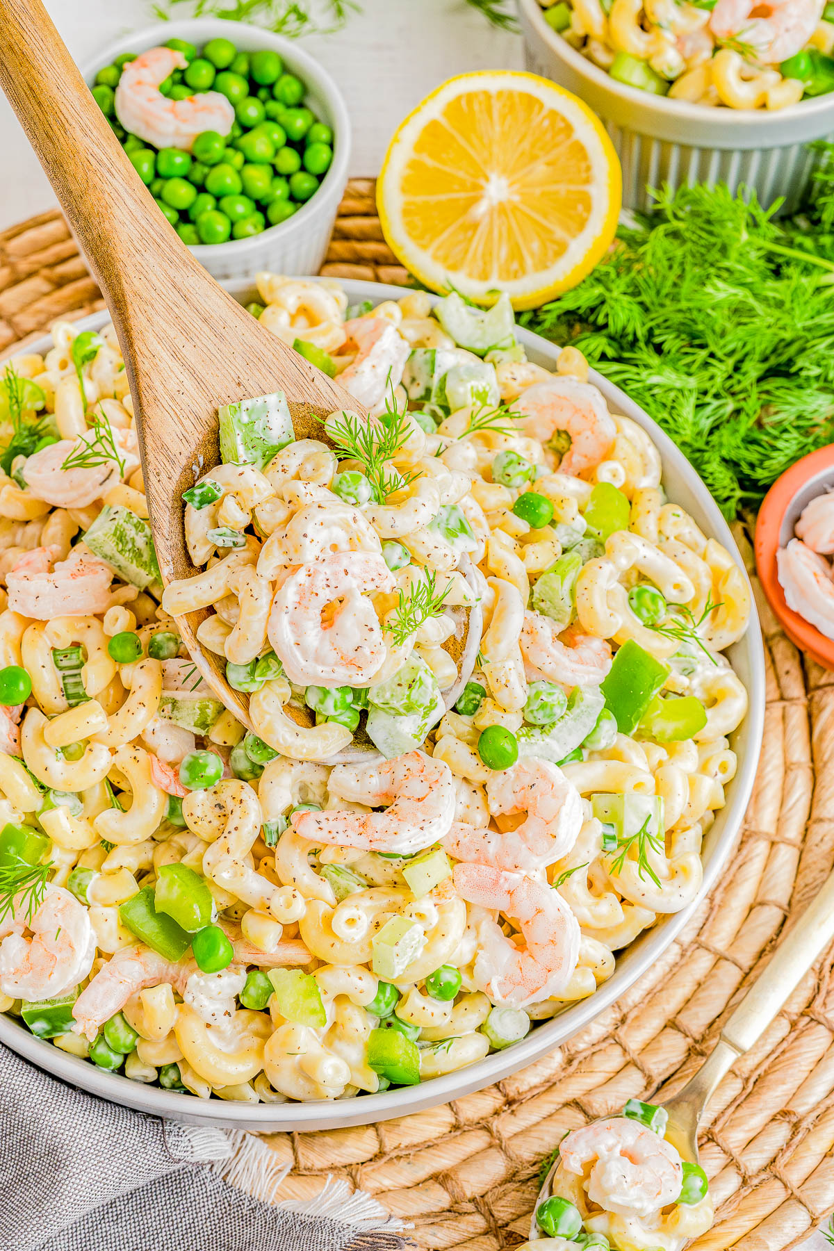 A vibrant bowl of shrimp pasta salad with peas, green peppers, and dill, served on a woven placemat with a wooden spoon. lemon half and bowl of peas in the background.
