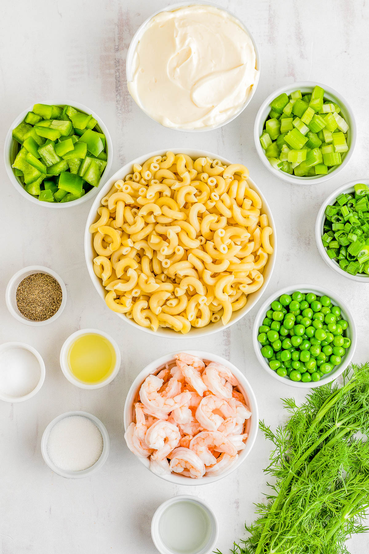Ingredients for making shrimp pasta salad displayed in small bowls, including cooked macaroni, shrimp, peas, chopped celery, and seasonings on a light surface.