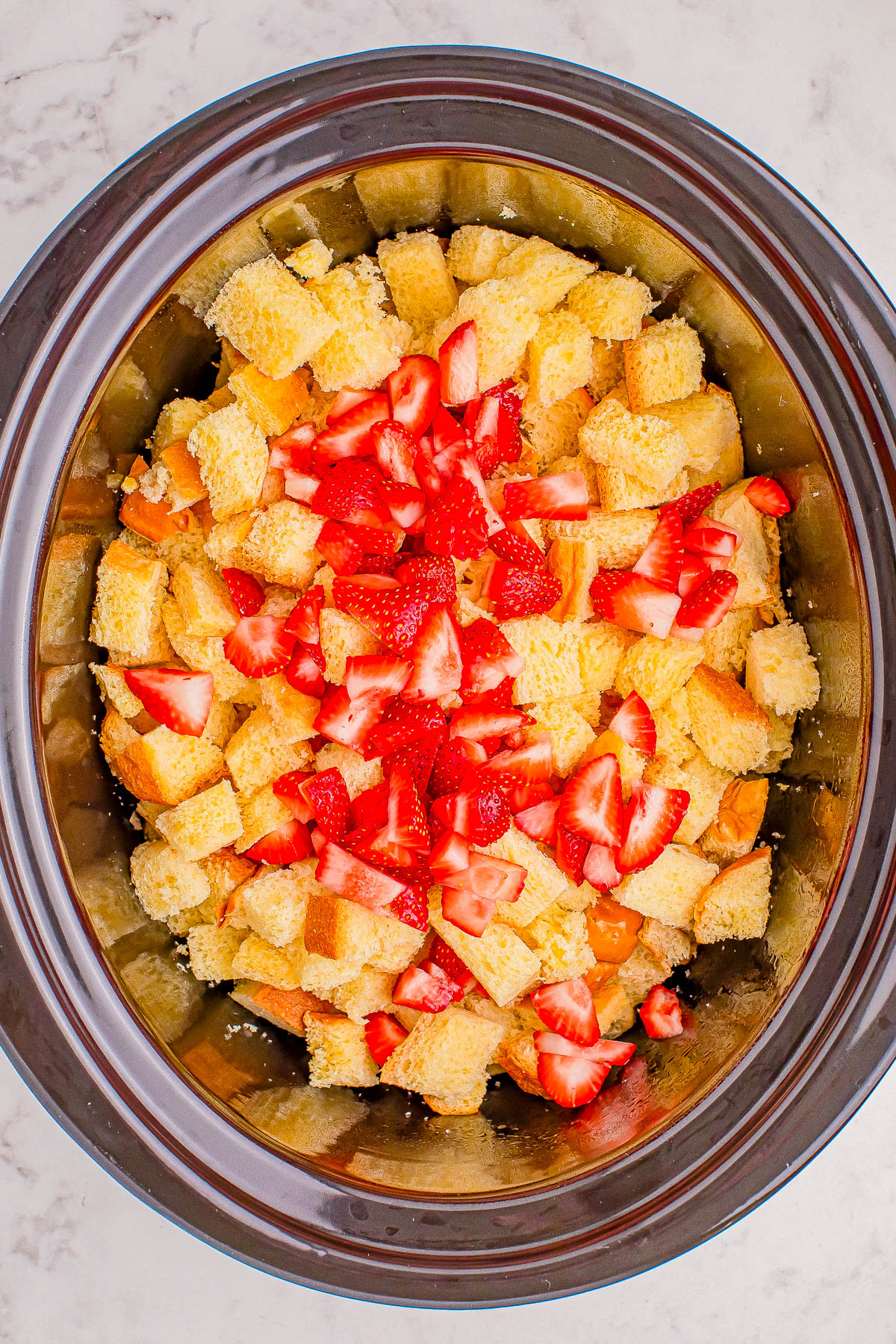 A slow cooker filled with cubed sponge cake and chopped strawberries on a marble surface.
