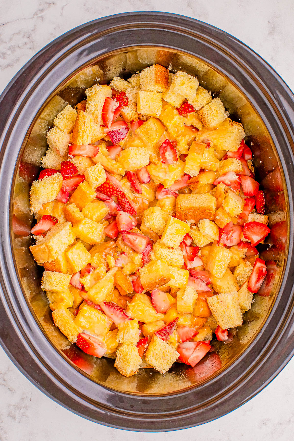 A slow cooker filled with a mixture of cubed bread, sliced strawberries, and peach chunks, set on a marble countertop.