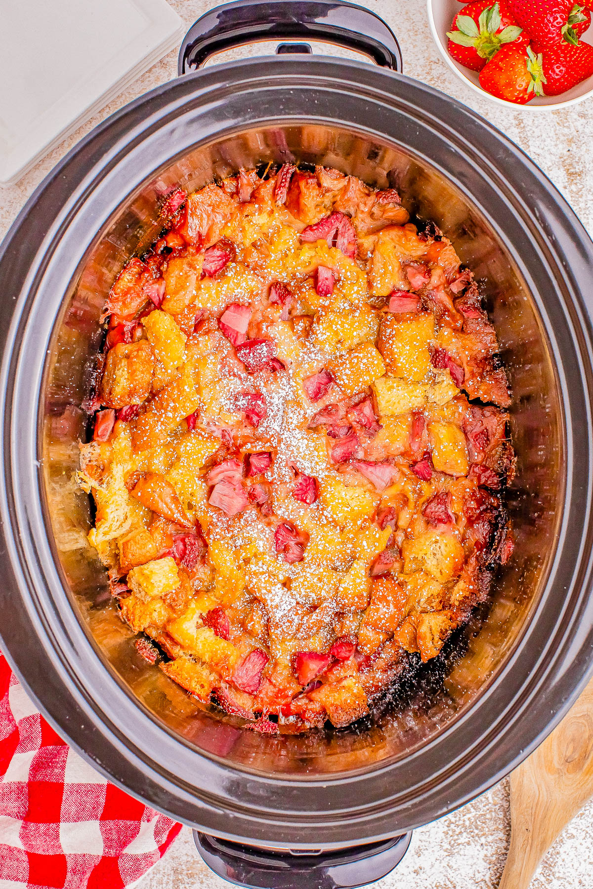 A cooked dish in a slow cooker, consisting of diced bread, chopped bacon, and melted cheese. A bowl of strawberries and a red checkered cloth are nearby.