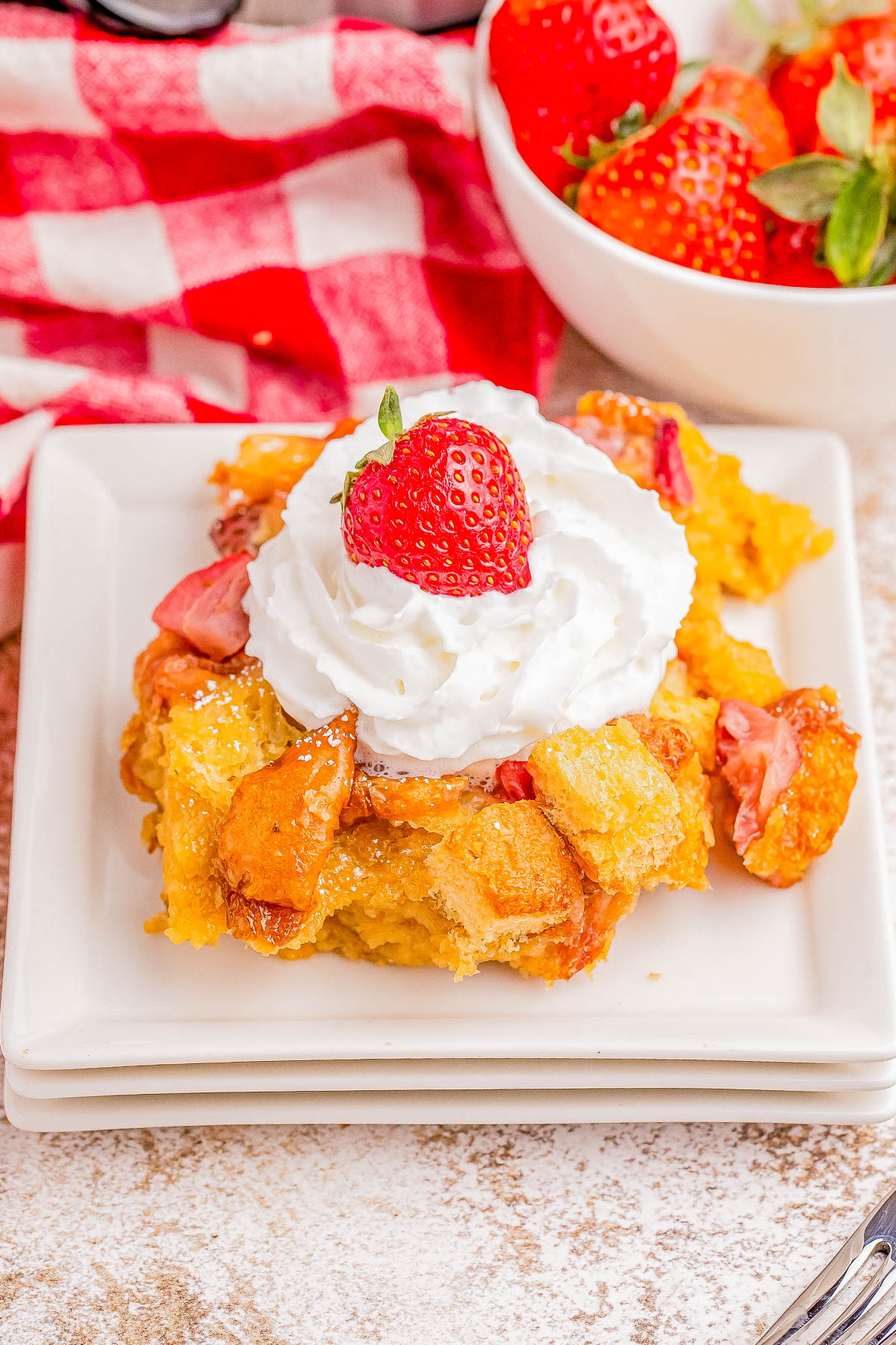 A dessert plate with a slice of bread pudding topped with whipped cream and a fresh strawberry. A bowl of strawberries and a red-and-white checkered cloth are in the background.