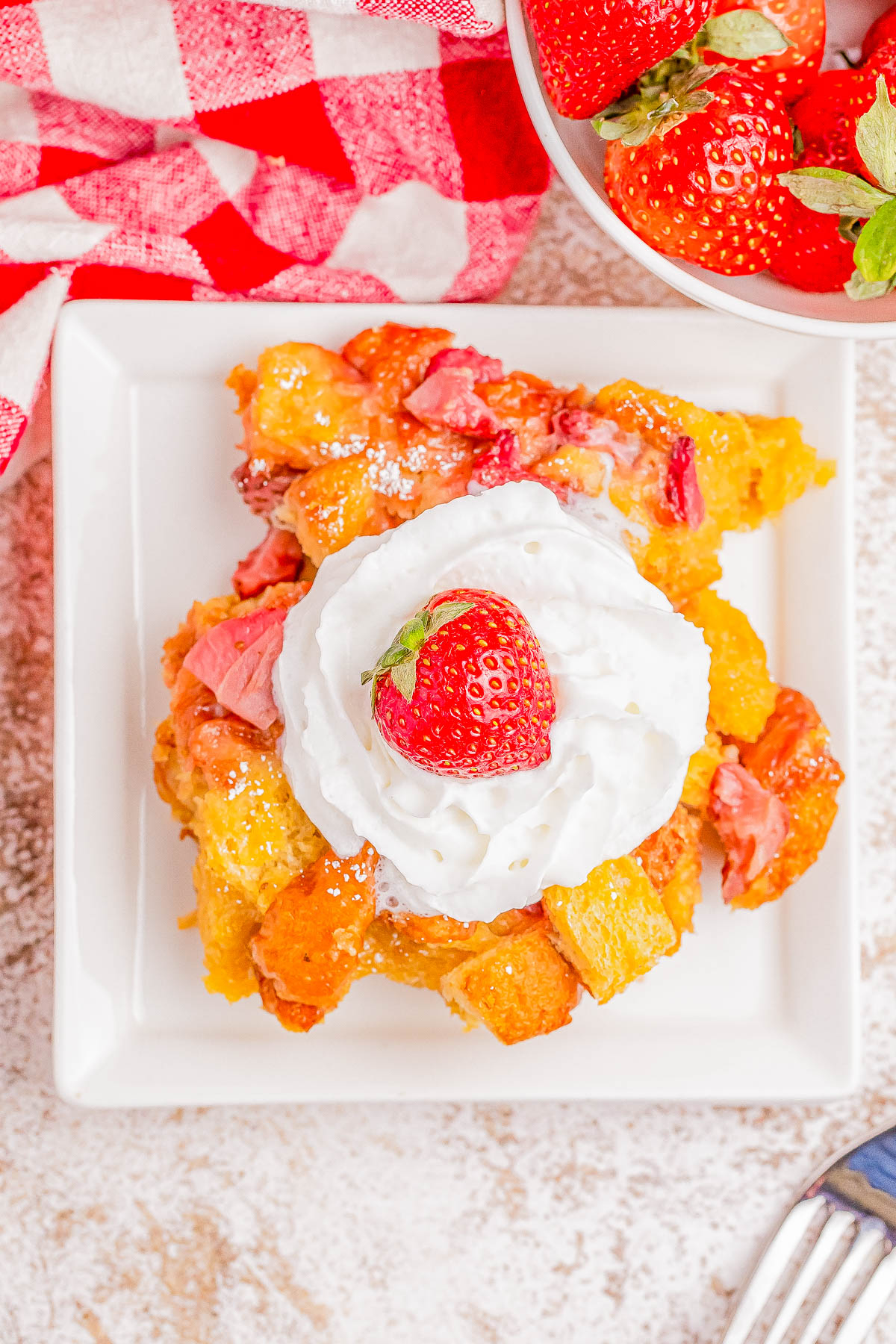 A square plate of strawberry bread pudding topped with whipped cream and a fresh strawberry, with a bowl of strawberries and a red-checkered cloth in the background.