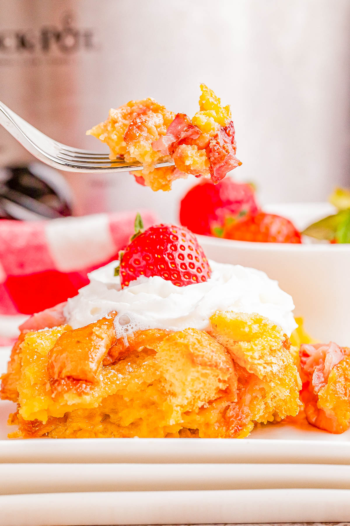A fork holds a bite of strawberry bread pudding with a slice of cake topped with whipped cream and a strawberry on a plate below.