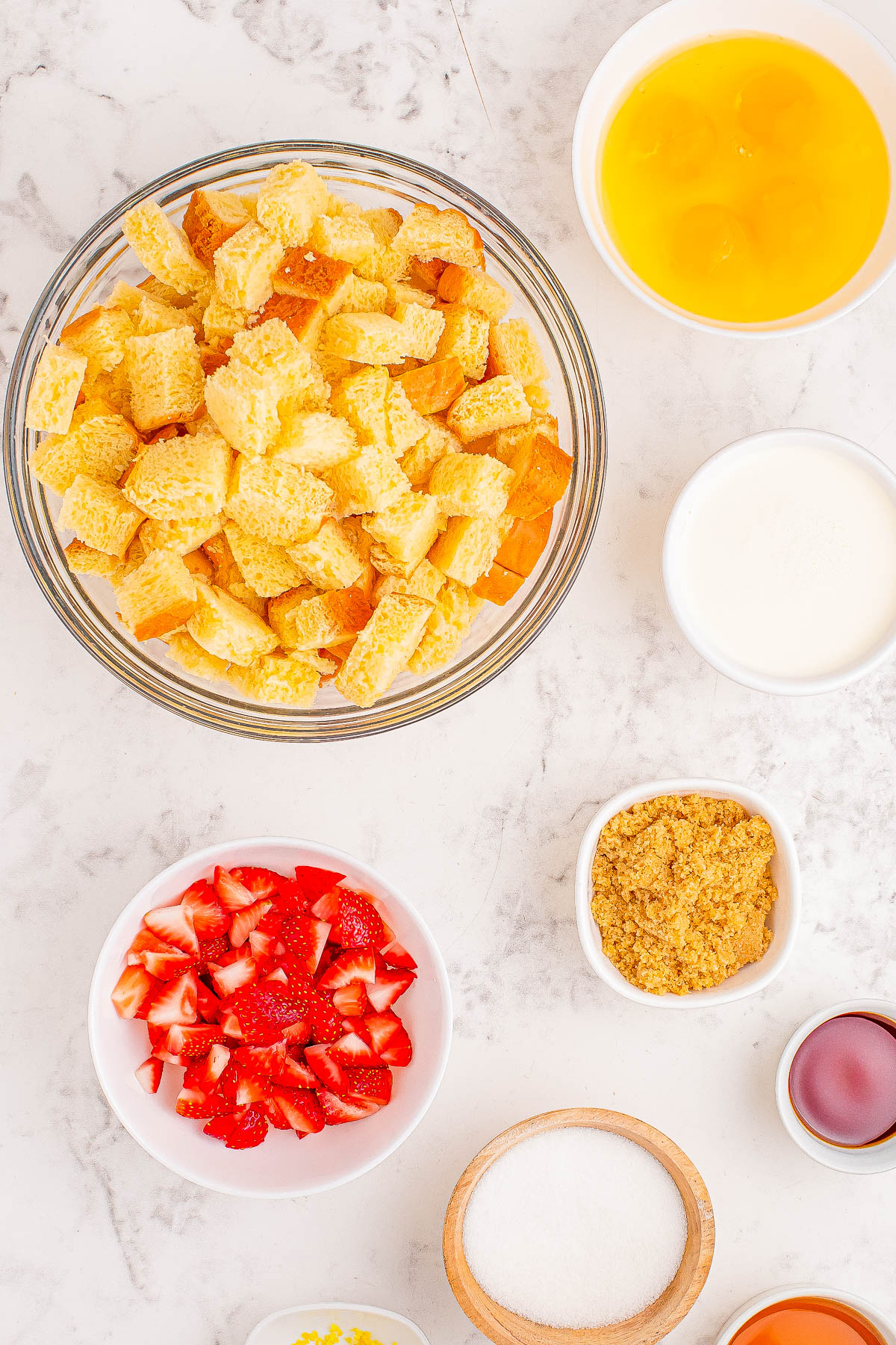 Ingredients for a recipe on a marble countertop include cubed bread, strawberries, eggs, brown sugar, granulated sugar, vanilla extract, milk, and lemon zest.