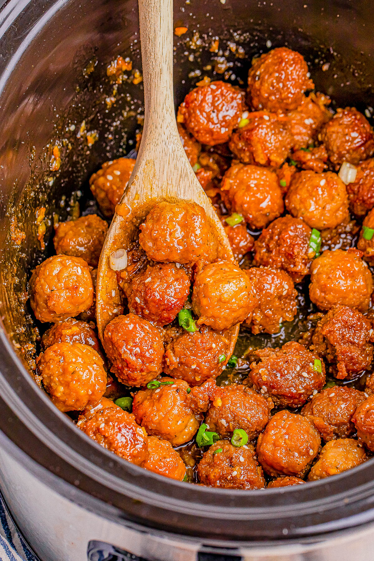 Wooden spoon stirring glazed meatballs in a slow cooker, garnished with chopped green onions.