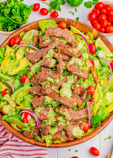 A wooden bowl filled with a salad featuring sliced steak, cherry tomatoes, avocado, red onion, and a green dressing. Other ingredients like parsley and lime are visible in the background.