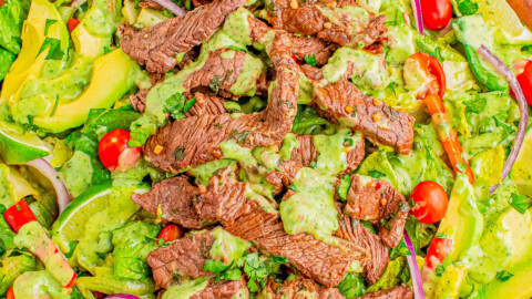 A wooden bowl filled with a salad featuring sliced steak, cherry tomatoes, avocado, red onion, and a green dressing. Other ingredients like parsley and lime are visible in the background.