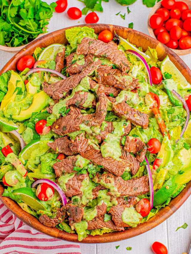 A wooden bowl filled with a salad featuring sliced steak, cherry tomatoes, avocado, red onion, and a green dressing. Other ingredients like parsley and lime are visible in the background.